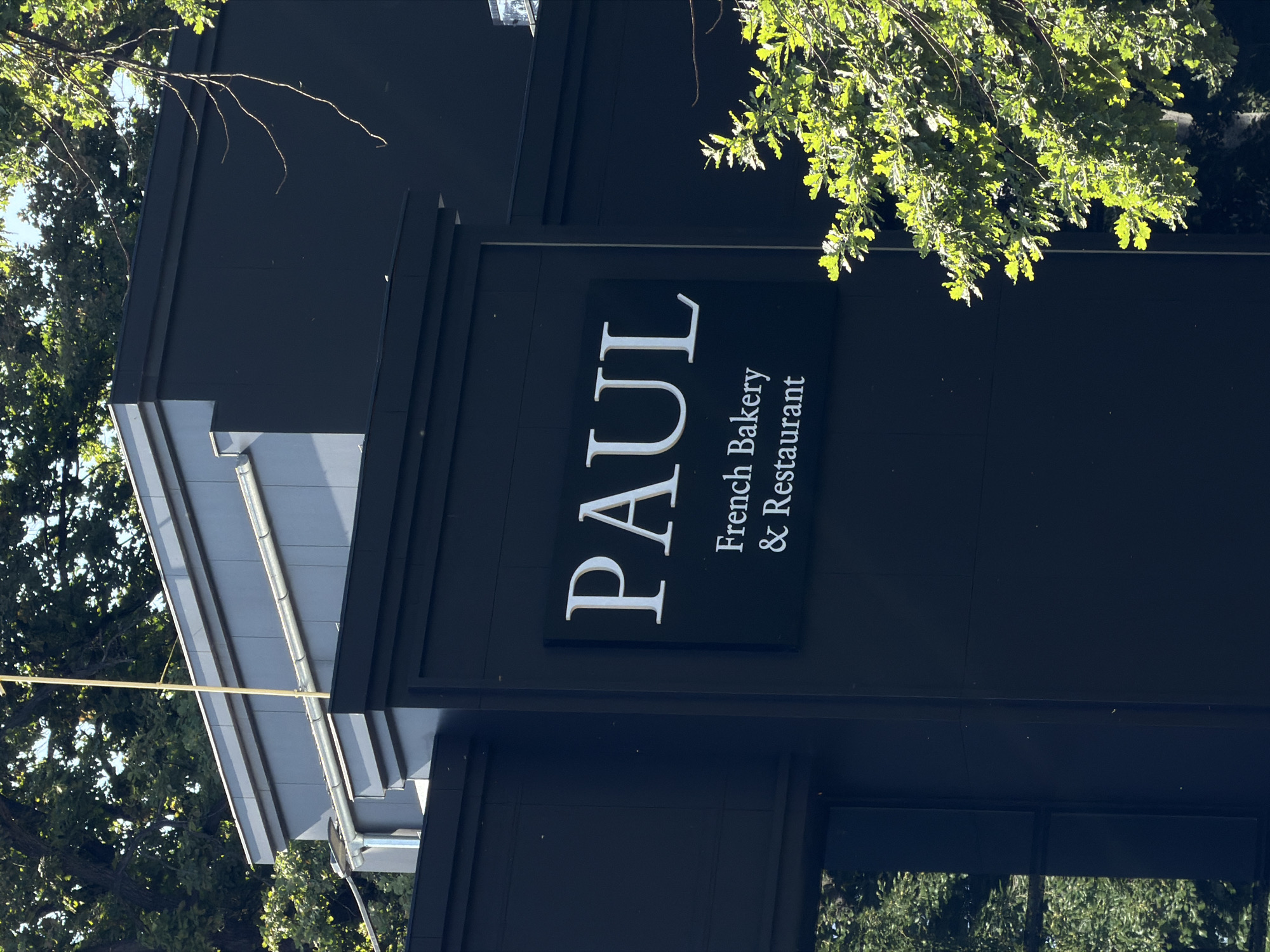 The image shows the exterior of a Paul bakery and restaurant, a French bakery chain. The restaurant appears to be located in Bishkek City, Kyrgyzstan. The building is dark-colored with a sign displaying the name "PAUL" in large, white letters, along with the words "French Bakery & Restaurant" beneath it. Some trees are visible in the foreground and background, partially obscuring the building. The photograph is taken at an angle, showing a section of the building's roof and the surrounding foliage.