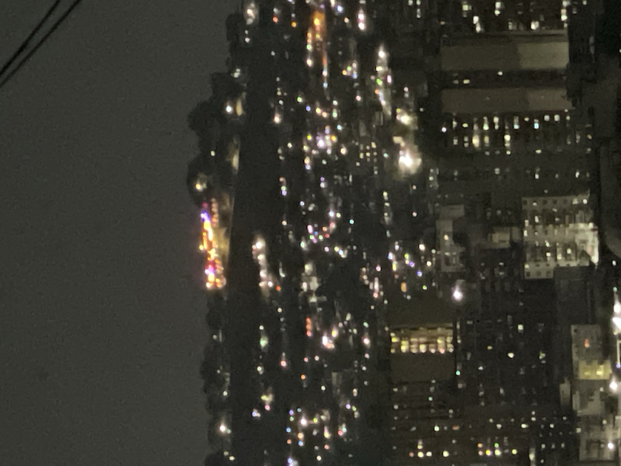 The image shows a blurry nighttime scene of buildings in Caracas, Venezuela. The buildings are tall and densely packed, with numerous windows illuminated, creating a glittering effect. The sky is dark and cloudy. A few overhead wires are visible in the upper left of the frame. The image is out of focus and lacks sharpness.