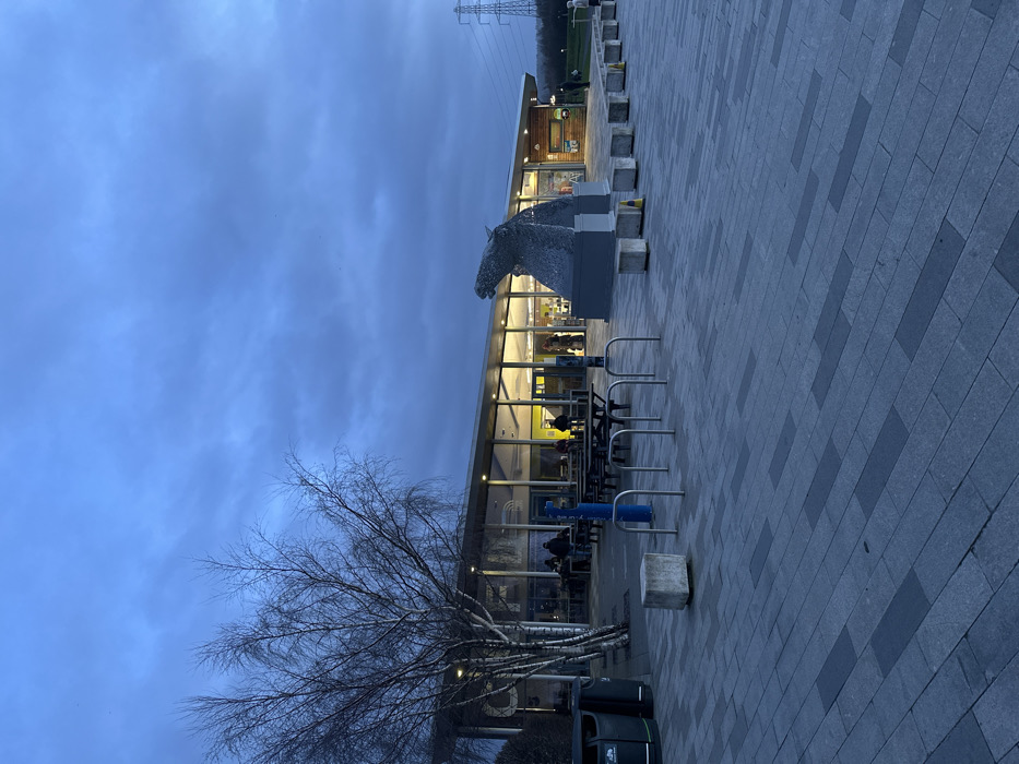 The image depicts an outdoor scene at dusk or early evening at the visitor center for The Helix park, located in Falkirk, United Kingdom. The sky is overcast and uniformly blue-grey.
The central subject is a modern, single-story building with an extensive glass-paneled facade and a sloped roof. The building's interior is brightly illuminated with warm yellow light, revealing approximately 10-15 individuals; some are seated at tables, while others stand near what appears to be a counter or service area. Directly in front of the building's main entrance stands a large, metallic sculpture of a horse's head, constructed from interwoven metal strips, resting on a grey rectangular plinth. This sculpture represents one of The Kelpies.
To the left of the building, a tall, bare-branched tree is visible, alongside several dark brown wooden picnic tables with integrated benches positioned on a paved area. Two dark green outdoor waste bins are located further to the left.
The foreground is a large paved plaza composed of rectangular grey and darker grey paving slabs arranged in a linear pattern. This area features blue metal bicycle racks and a series of light grey concrete block bollards or seating elements.
In the distant background to the right, a grassy expanse leads to a line of trees, and a prominent metal electricity pylon is visible against the sky. A few indistinct human figures are also present in this distant right background.