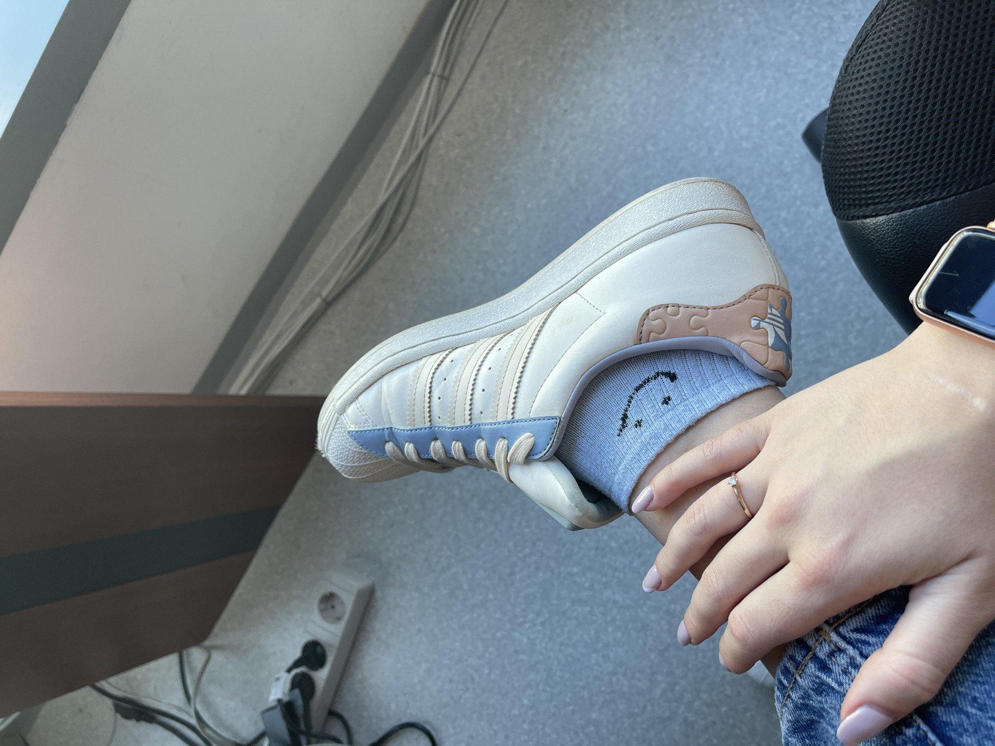 A medium-angle shot depicts a person's left foot clad in an Adidas sneaker with a blue toe, cream-colored body, and light brown accents. The shoe is worn with a gray sock featuring a black smiley face. The person's hand, adorned with a gold ring, rests on the ankle. The person wears denim jeans and an Apple Watch on their wrist. A black office chair is partially visible in the top right corner. In the background, there is a wall with an electrical outlet and cables, along with a window. The setting could be an office or home in Sofia, Bulgaria, but this cannot be conclusively determined from the image.