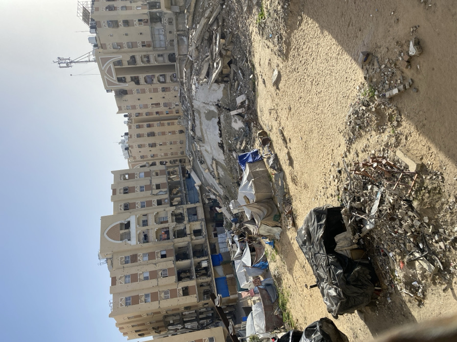 The image depicts a scene of extensive urban destruction and makeshift habitation in Khan Yunis, Palestinian Territory, under clear daylight. In the background, multiple multi-story residential buildings, primarily tan-colored, show significant damage, including numerous holes on their facades consistent with ballistic impacts or shrapnel, shattered windows, and collapsed or gutted sections on lower floors. Distinctive arched architectural features are visible on some buildings, and antennae towers are present on rooftops.
In the midground, a large mound of concrete rubble, twisted rebar, and debris dominates the center-right, indicating the complete collapse of a substantial structure. To the left, on sandy ground, a cluster of approximately 10-15 rudimentary shelters is established. These makeshift tents are constructed from various materials, including white, blue, and beige tarpaulins, blankets, and possibly salvaged materials, with some laundry visible hanging nearby.
The immediate foreground features sandy earth sparsely interspersed with small patches of vegetation. On the bottom left, a large pile of dark material, seemingly covered with black plastic sheeting, is present next to smaller accumulations of shattered concrete, twisted metal fragments, and general refuse. No individuals are visible within the frame.