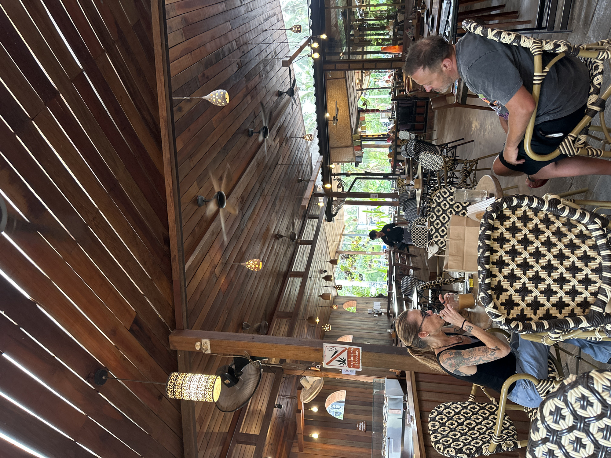 This is an indoor scene at a restaurant in Tambon Ko Pha Ngan, Thailand, with a rustic, wooden interior and an open-air feel. The ceiling is constructed from parallel wooden planks with gaps between them, allowing natural light to filter through, and is further illuminated by multiple hanging pendant lights and ceiling fans. In the foreground, a woman with blonde hair pulled back in a ponytail and wearing a black tank top and light blue pants is seated at a table, drinking from a glass with a straw. She has tattoos on her arms. To her right, a man with short, light brown hair and wearing a gray t-shirt and black shorts is seated, looking towards his left. Both are seated in chairs with woven rattan frames and patterned upholstery. A sign that reads "NO SMOKING CANNABIS" is visible on a wooden post in the mid-ground, next to a large industrial fan. The background shows more tables and chairs, with patrons and staff visible in the distance, and lush green foliage through the open sides of the restaurant. The overall atmosphere suggests a casual dining establishment.