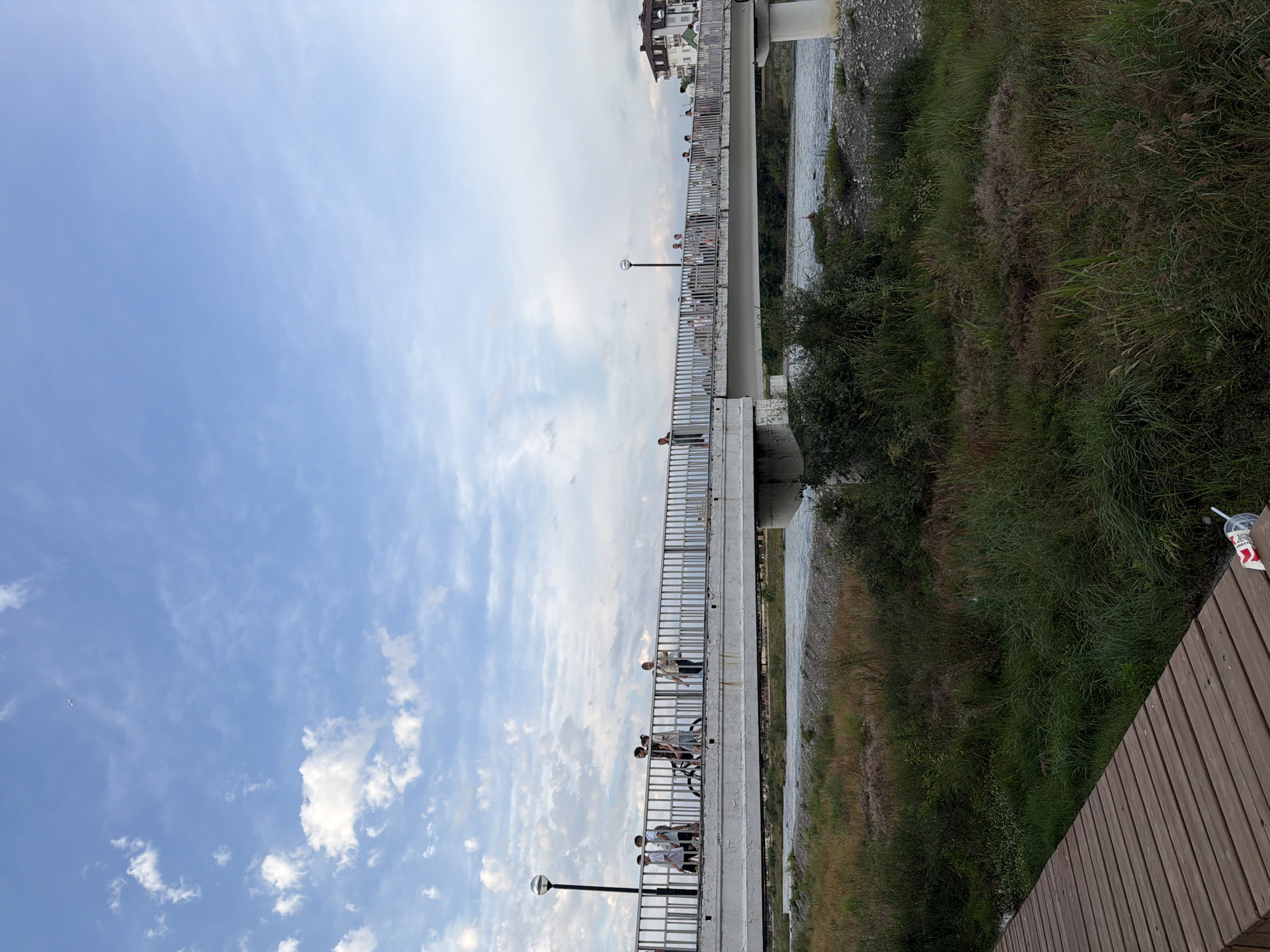 A long, pedestrian bridge crosses a river in Sochi, Russia. The bridge has a metal railing and is lined with people. Some individuals are standing and looking out at the river, while others are seated or have bicycles with them. The sky is partly cloudy with patches of blue. The river flows beside a grassy embankment. On the right side of the image, a wooden boardwalk is visible, with a cup and straw on it. The time of day appears to be late afternoon or early evening. The overall mood is casual and relaxed, with people enjoying the outdoors.