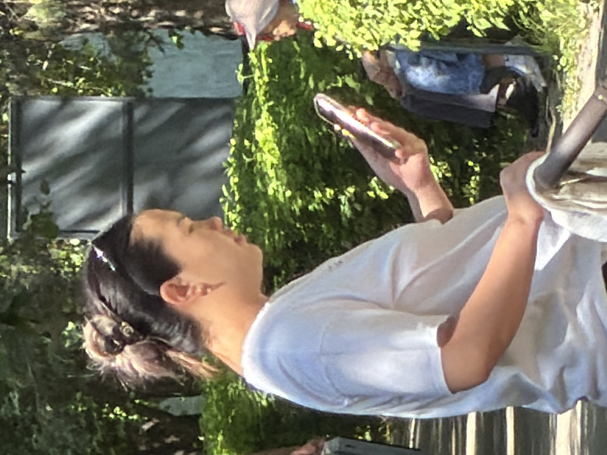 A woman with dark hair pulled into a messy bun, adorned with a decorative hair clip, is shown in profile, looking down at her phone. She is wearing a white t-shirt and a small earring in her left ear. Her right hand holds the phone, which is silver and appears to have a light-colored case. Behind her, partially obscured by lush green bushes, an elderly person wearing a hat and denim jeans is visible. The scene takes place outdoors in an area with trees and foliage, suggesting a park or garden setting. The lighting indicates it is daytime, with sunlight filtering through the leaves. A dark-colored pipe or railing is in the foreground, with a light-colored cloth draped over it.