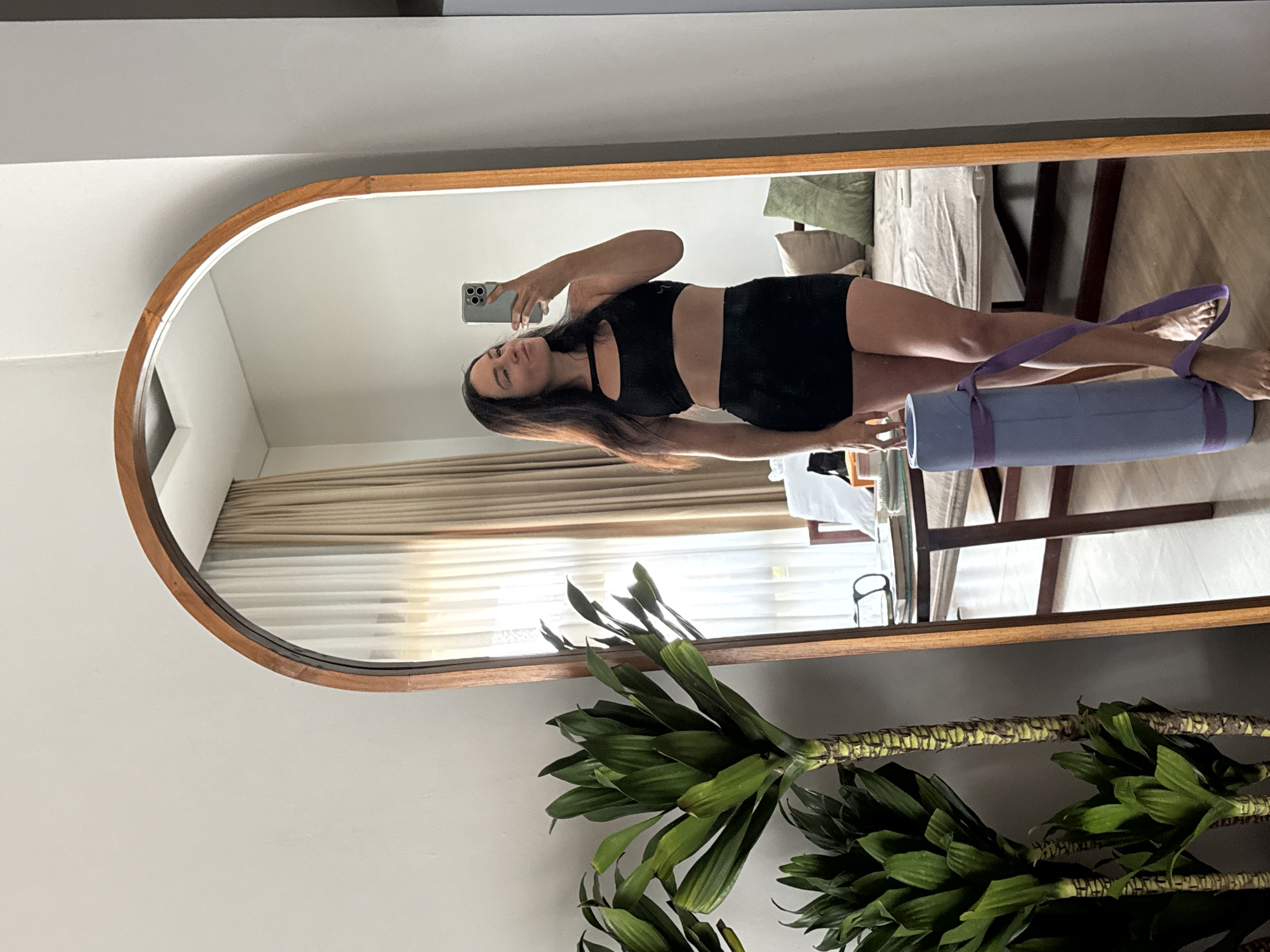 A full-body mirror reflects a woman with dark hair, wearing a black sports bra and black shorts. She is holding a rolled-up yoga mat with purple straps in her left hand and a smartphone in her right hand, taking a selfie. She is standing in what appears to be a bedroom in North Kuta, Indonesia, with a large potted plant to her left. In the reflection, a bed with beige bedding and green pillows is visible behind her, along with a wooden table with books and a pair of glasses. Light brown curtains hang in the background, suggesting the room receives natural light.