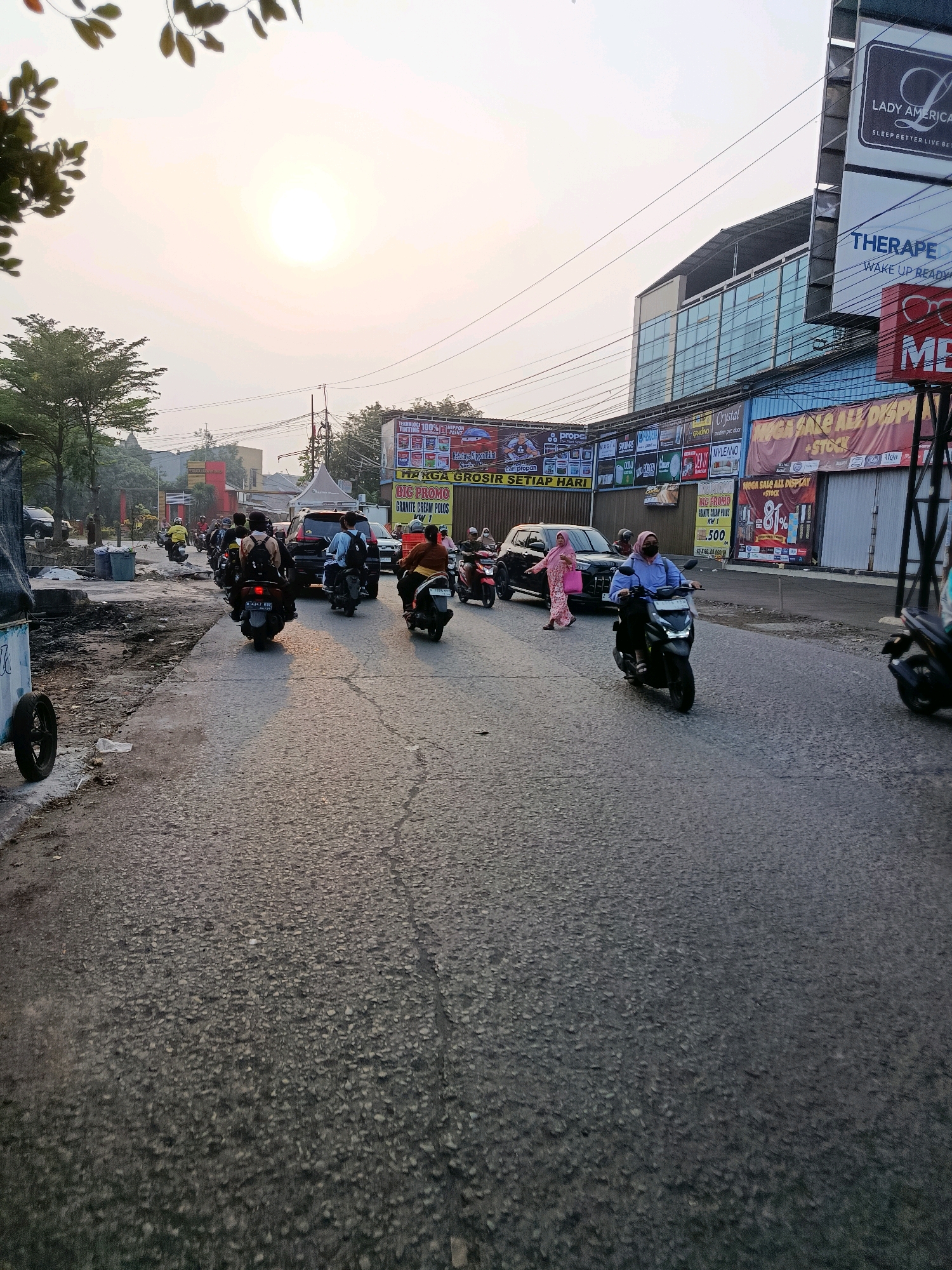 The image depicts a street scene in Bekasi, Indonesia, during what appears to be late afternoon or early evening, with the sun low in the sky. The road is paved and shows signs of wear. A line of motorbikes is moving along the road, with several riders wearing helmets. One motorbike in the foreground has a black body and a rider in a blue shirt and a mask. To the right of this motorbike, a woman in a pink headscarf and dress is crossing the road in front of a black car. Further down the road, more motorbikes and cars are visible, indicating moderate traffic.
Along the right side of the street, there are several buildings with signs. One prominent sign displays "LADY AMERICA" and "THERAPE WAKE UP READY". Another large sign has bold lettering, including "MEGA SALE ALL DISPLAY + STOCK" and a percentage figure "81%". Other signs indicate promotions for paint products and various brands. The buildings appear to be commercial establishments.
On the left side of the road, there is a construction or debris area with piles of dirt and rubble. Some trees are visible in the background, and the overall atmosphere suggests an urban environment in Indonesia. The lighting conditions indicate a golden hour setting.
