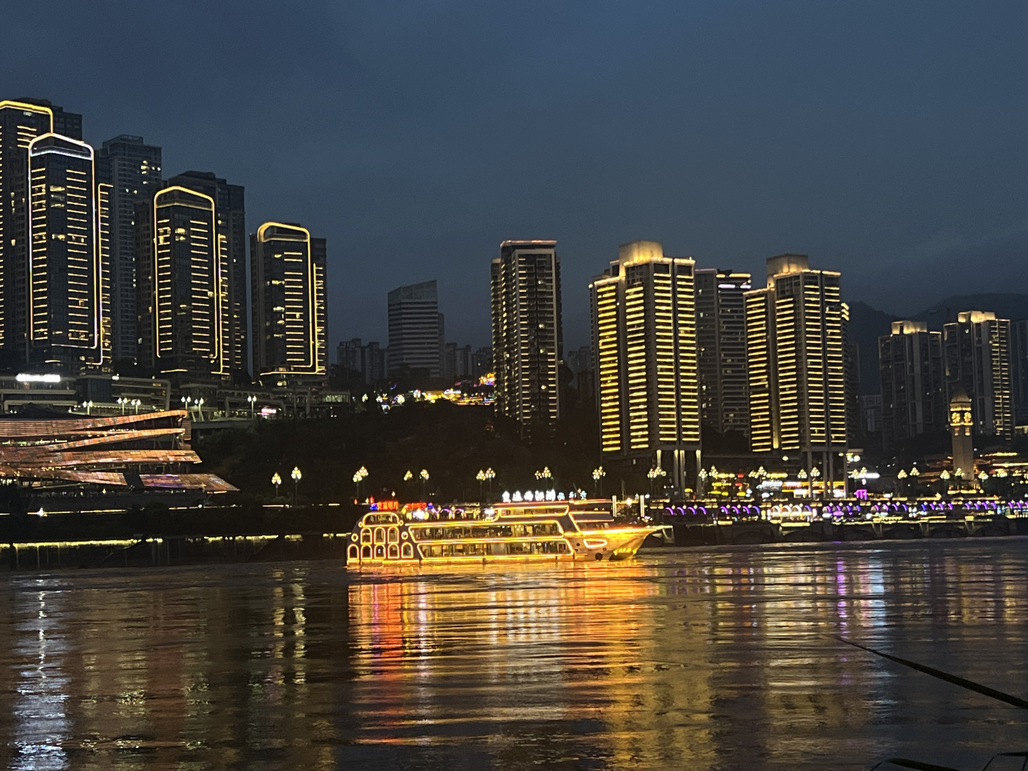 This image captures a nighttime scene along the riverfront in Jiangbeicheng Subdistrict, Chongqing, China. The foreground features the calm, reflective surface of a wide river, illuminated by the bright lights of the city. A large, multi-deck passenger boat, adorned with numerous yellow and orange lights, sails across the water. The boat is the primary focal point in the river.
In the background, the Jiangbeicheng cityscape rises, characterized by a dense cluster of modern, illuminated skyscrapers. Many of these buildings are outlined with bright yellow lights, highlighting their vertical structures against the dark twilight sky. To the left, a layered structure with illuminated orange and pink lights is visible, possibly a modern building or entertainment complex. A prominent clock tower with illuminated features stands out further down the riverfront. The overall atmosphere is one of urban vibrancy at night, with the city's lights creating striking reflections on the water. There are no discernible people in the image.