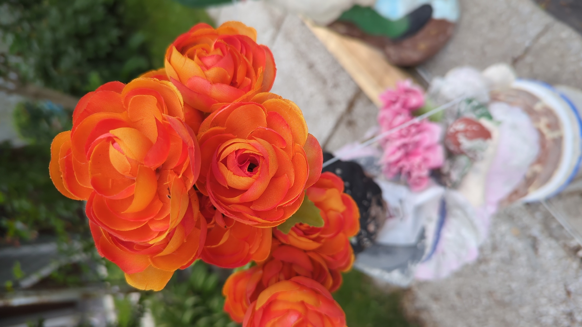 The image shows a close-up of a bouquet of artificial orange flowers in a planter. The flowers are vibrant and have many petals. In the background, there is greenery and a blurred view of what appears to be a patio or outdoor area. The planter itself is partially visible, containing some decorative items. The overall scene is focused on the floral arrangement, suggesting it might be part of a garden or outdoor decoration. The image does not contain any information that would indicate its location in Montreal, Canada.