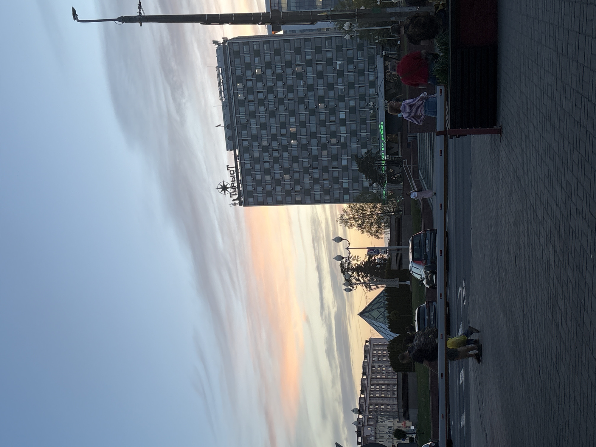 The image shows an urban scene in Minsk, Belarus, during a sunset. Prominent is the "Tourist" hotel, a tall, rectangular building with a sign on top. The sky is filled with striated clouds tinged with pink and orange from the setting sun. A construction crane is visible in the upper portion of the photo. In the lower part of the image, a paved area is visible with people present. A low barrier, possibly for controlling access, spans the width of the frame. Cars are parked beyond the barrier. People are standing and kneeling near the barrier.