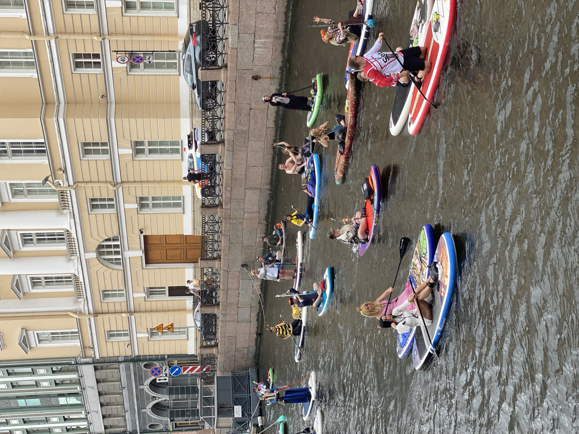 This image shows a group of people paddleboarding on a canal in Saint Petersburg, Russia. The buildings in the background are characteristic of the city's historic architecture, with ornate facades and classical detailing. The canal appears to be lined with stone walls and a decorative fence. Several people are dressed in costumes, suggesting a themed event. The water is a murky brown, and the sky is overcast. The paddleboarders are of various ages and genders, and they are all holding paddles. Some are sitting, while others are kneeling or standing. The scene is lively, with many participants and onlookers visible.