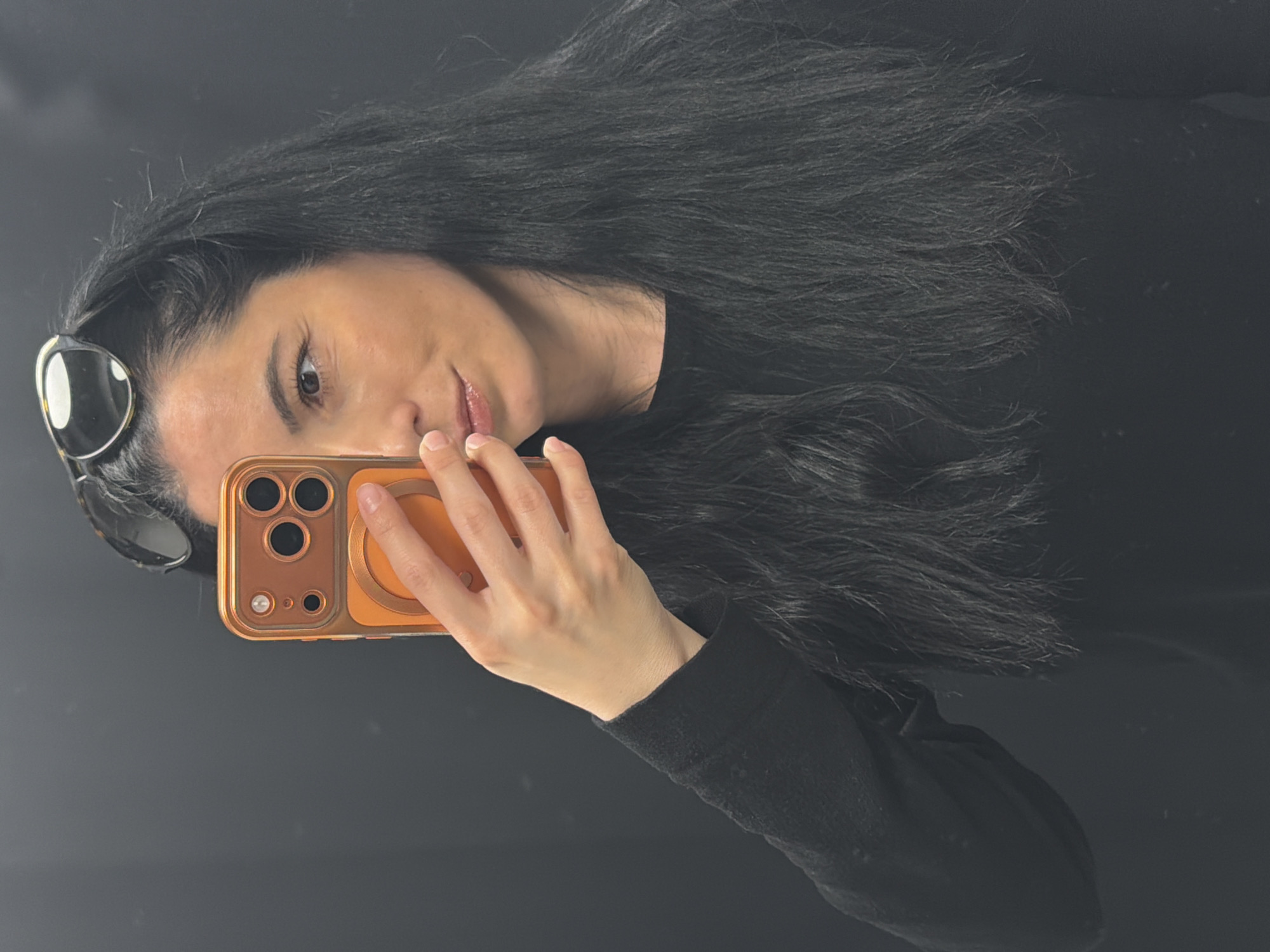 A woman with long, black hair is holding an orange mobile phone with three camera lenses in front of her face. She is wearing sunglasses pushed up on her forehead. She is wearing a black long-sleeved shirt. The background is a dark gray or black. There is no indication of the location being Pleven, Bulgaria.