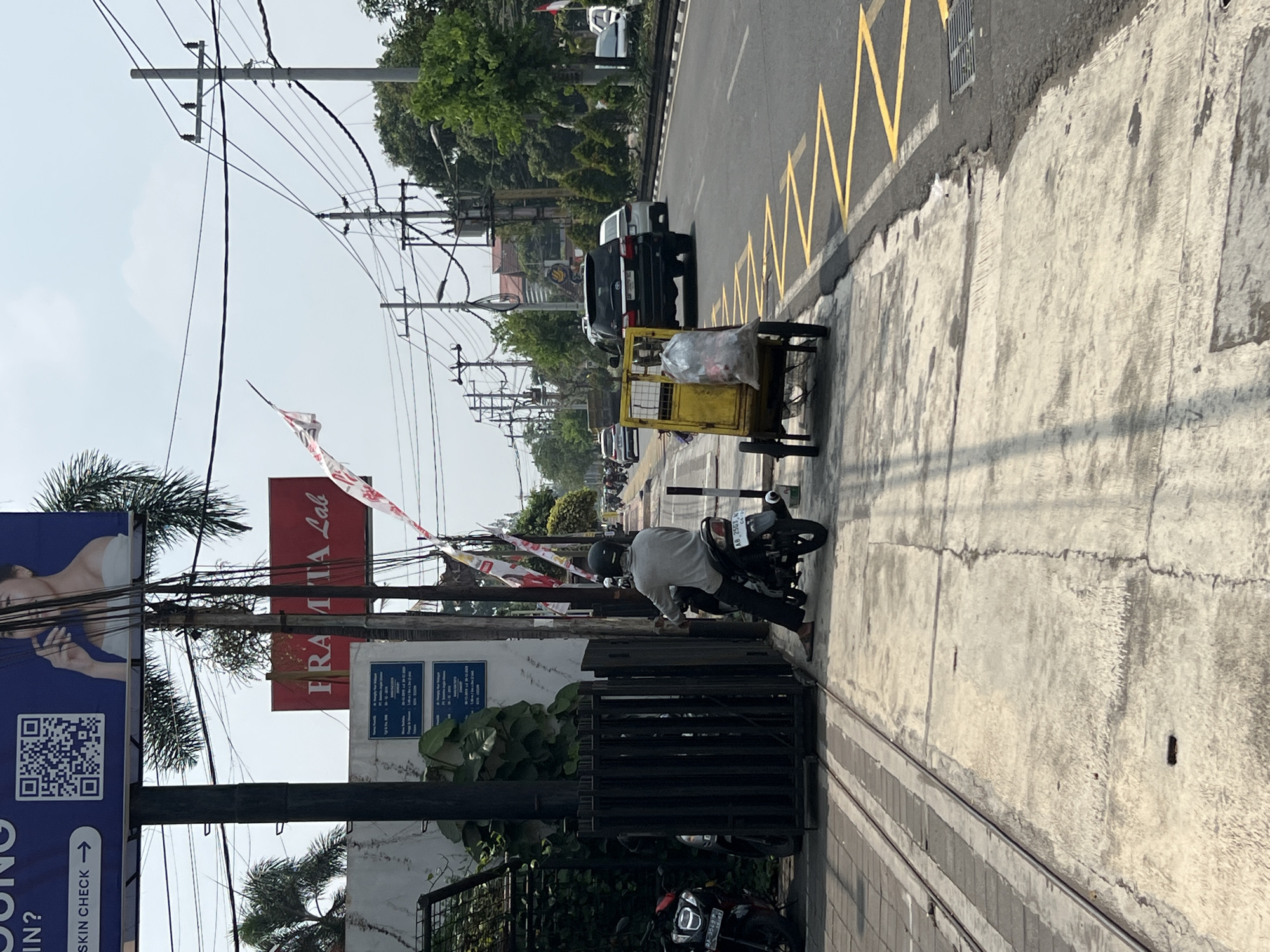 The image captures a street scene in Yogyakarta, Indonesia, on a bright, sunny day. A person is seated on a motorcycle, wearing a grey long-sleeved shirt and dark pants, with a black helmet. They are positioned on a sidewalk next to a yellow cart containing a large plastic bag. The sidewalk is made of concrete and has visible cracks. Adjacent to the sidewalk are train tracks.
In the background, a busy road features several vehicles, including a black SUV, a white sedan, and other cars and motorcycles. Trees line the side of the road, and utility poles with numerous wires stretch across the sky. Above the road, billboards are visible. One prominent red billboard reads "FRAMITA Lab." Another large blue billboard displays a QR code and text that includes "SKIN CHECK →". The overall environment suggests an urban setting with ongoing street activity.