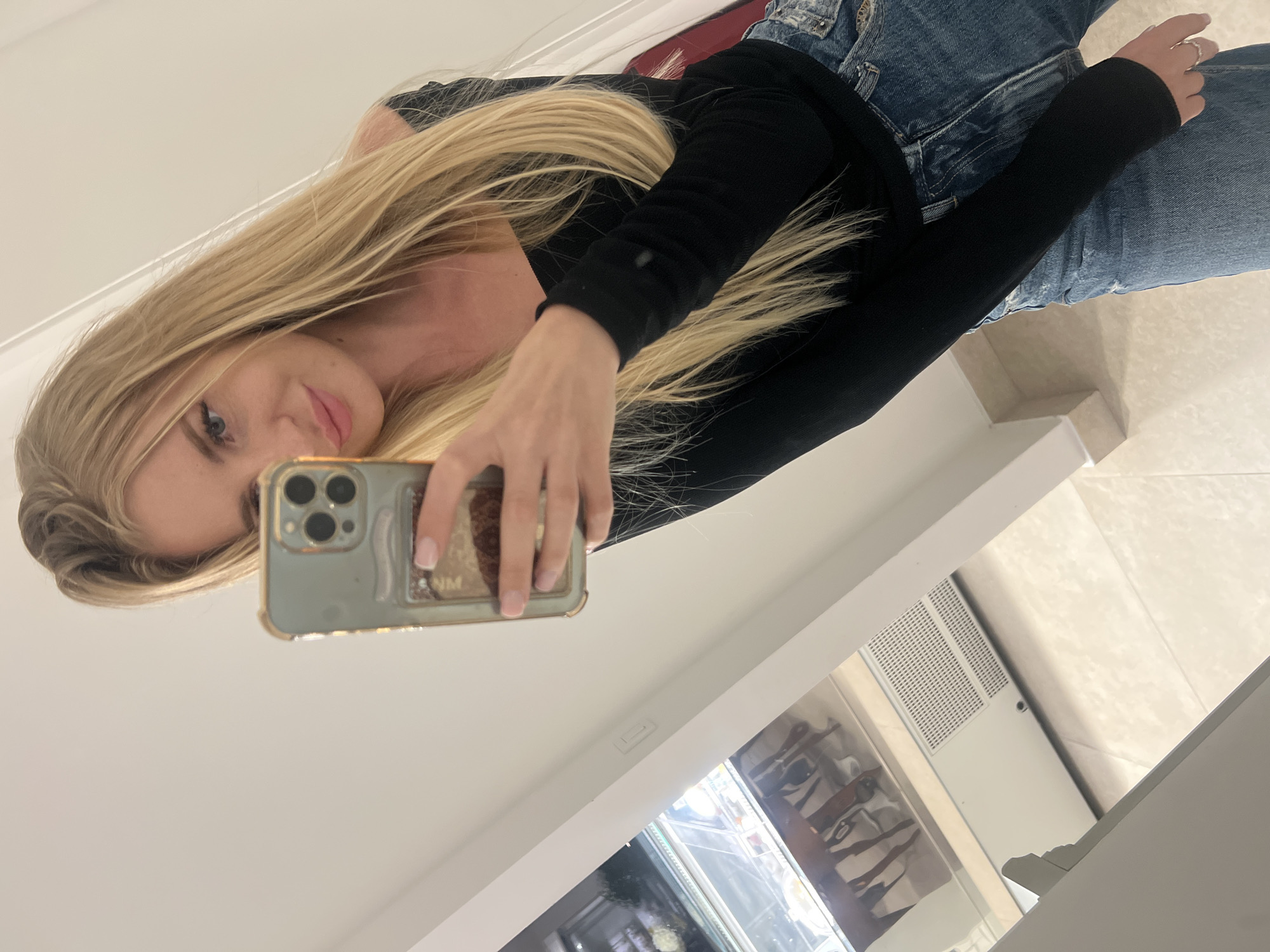 A woman with long blonde hair is taking a selfie in a mirror. She is wearing a black long-sleeved shirt and blue jeans. The mirror reflects a display case with various items inside. Based on the architecture and general style, the location is possibly within a business in the city of Bečići, Montenegro.