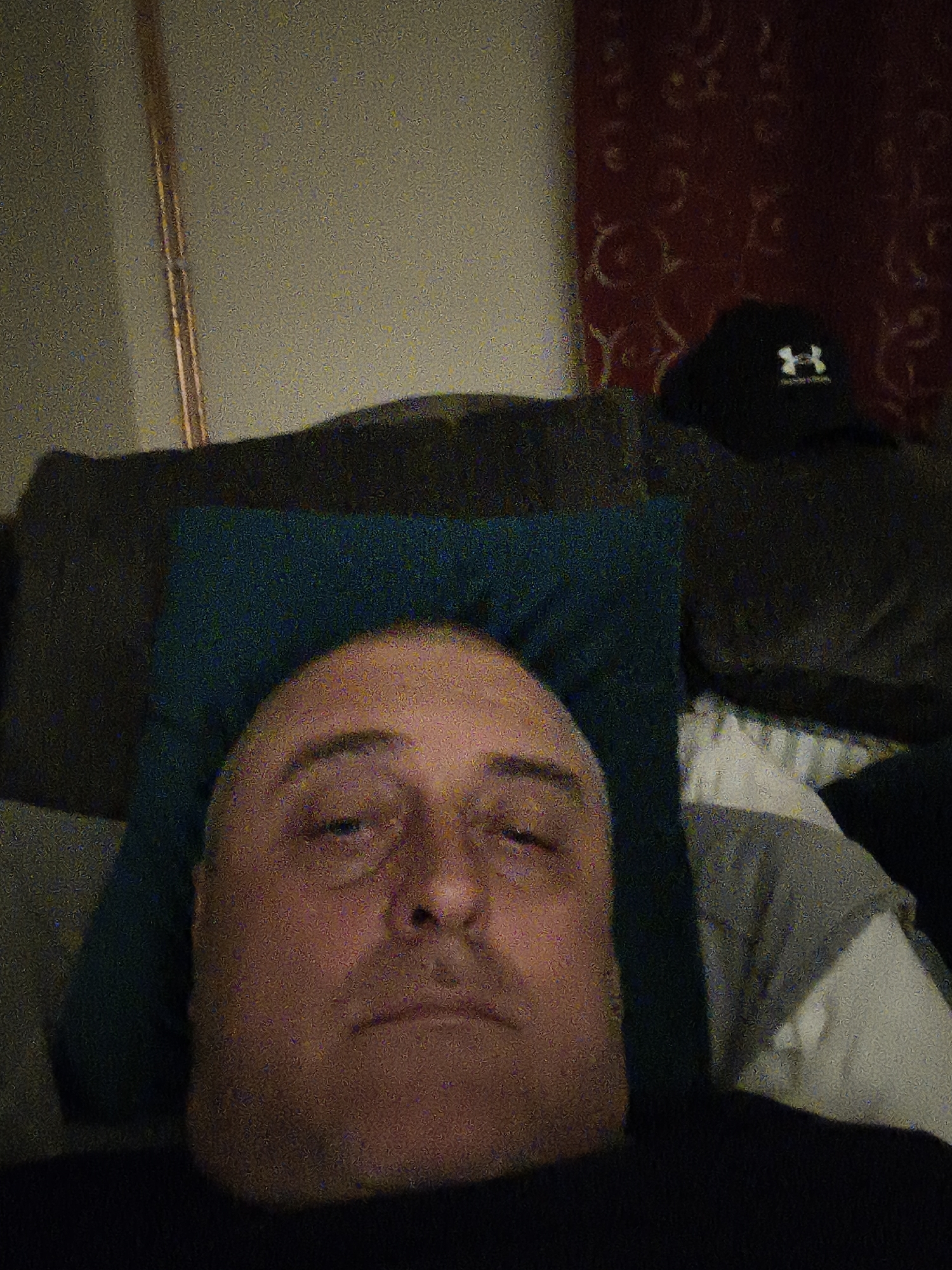 The image captures a middle-aged man lying down, resting on a dark blue pillow. His head is centered in the frame, and he is looking directly towards the viewer with a neutral, slightly tired expression, his eyelids appearing heavy. He is wearing a dark-colored top, possibly a t-shirt.
The setting appears to be indoors, likely a bedroom or living room, suggested by the presence of a couch or bed, pillows, and decorative elements. Behind the man's head, there is a dark-colored couch or headboard. To the upper right, a black baseball cap with a visible white "Under Armour" logo is placed on the furniture. The background on the right features a dark red wall or fabric with a subtle, lighter-colored intricate pattern. On the far left, a slender, vertical, metallic or copper-toned pipe or rod is visible against a plain, lighter-colored wall. A white sheet or blanket is partially visible to the man's right.
The overall lighting is dim, suggesting it might be evening, night, or early morning. The image quality is notably grainy and noisy, characteristic of photography taken in low-light conditions. There are no direct cues about the weather. The scene depicts a quiet, relaxed moment. Given the provided context, the location of this scene is Huntingdonshire, United Kingdom.
