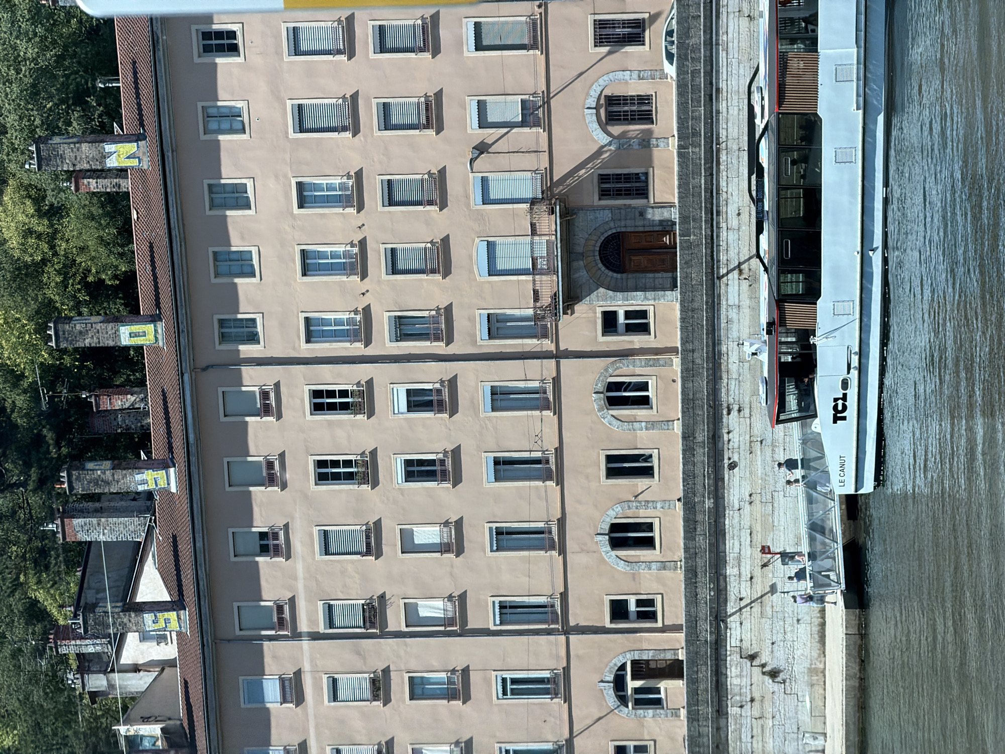 This is an aerial, slightly tilted, eye-level shot of a building facade and a riverboat on a river in Lyon, France. The building is multi-story with many windows, some with closed blinds and some with open shutters. The facade is a light beige color, and the roof is tiled. To the left of the building, trees are visible.
On the right side of the image, a white riverboat is docked. The boat has "TCL LE CANUT" printed on its side. Several people are visible on the boat, standing on a covered walkway and on the deck. The river is a murky brown color, and the day appears to be sunny, casting shadows from the building and its balconies. The scene is peaceful, with no discernible action beyond the presence of people on the boat.