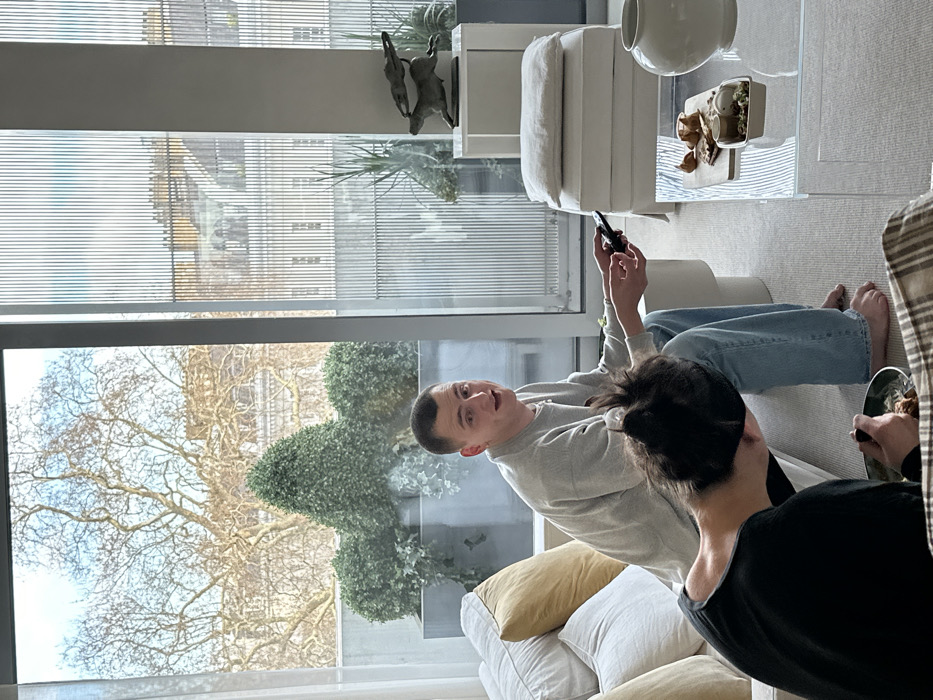 The image displays an indoor scene within a modern living room of an upscale residential property in London, United Kingdom, likely situated in a West or Central London area overlooking a garden square or park. Through large floor-to-ceiling windows, bare mature trees and classical stone buildings are visible. On an outdoor terrace, several rectangular planters contain trimmed topiary and trailing ivy.
Inside, two individuals are present. A male, with short hair and wearing a light grey long-sleeved top and light blue jeans, is seated barefoot on a white modular sofa. He holds a black remote control in his right hand and looks directly towards the camera. Partially visible in the foreground, with their back to the camera, is another individual with dark hair, wearing a black top. This person is leaning forward, eating from a green bowl with a fork.
A transparent rectangular coffee table is positioned in front of the sofa, holding a wooden serving board with various pastries, a small rectangular container of food, and another smaller container. A large white ceramic vase rests near the food items. To the right, a white pedestal displays a bronze sculpture depicting two running hares. The room features light-colored walls and a cream carpet.