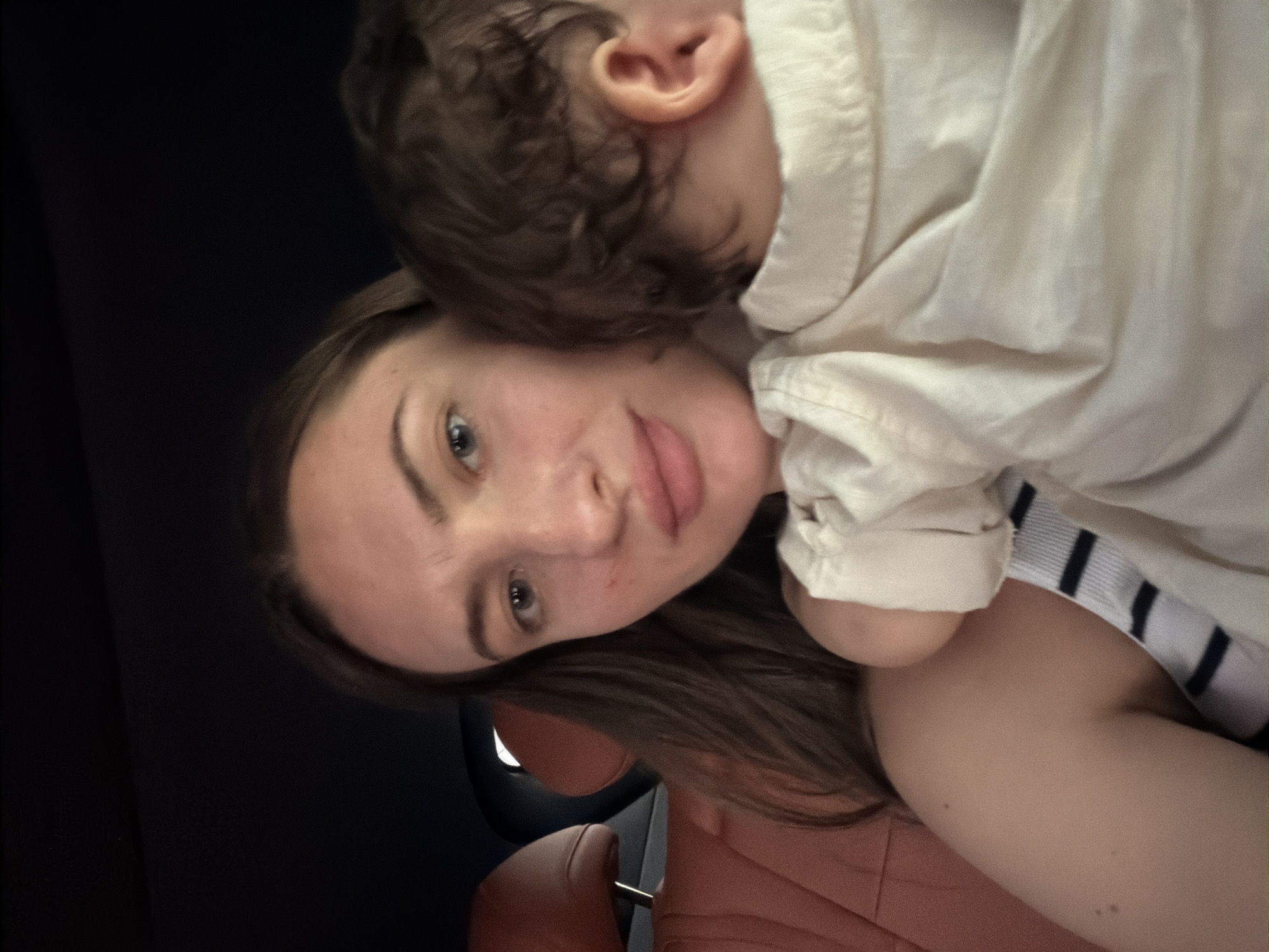 A close-up, selfie-style photograph shows a woman holding a baby in her arms. The woman has long, dark brown hair and light blue eyes. She is wearing a striped top. The baby is facing away from the camera, with its head resting on the woman's shoulder. The baby has dark, curly hair and is wearing a light-colored shirt. The background of the image is dark, but suggests the interior of a vehicle with a reddish-brown leather seat visible. The lighting is soft and natural. The image appears to have been taken indoors, possibly in a car. There are no specific landmarks or visual cues that place the image in Sharm El Sheikh, Egypt.