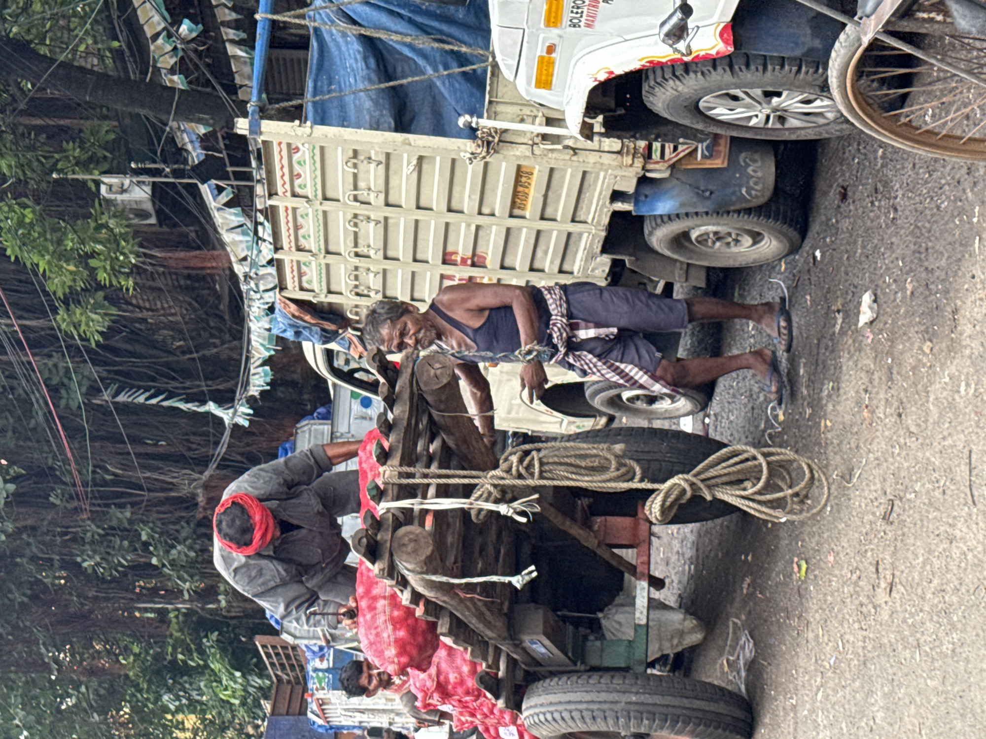 A street scene in Kolkata, India, features a man in a gray shirt and red headscarf bent over a cart loaded with red produce, likely onions, contained in netting. The cart, made of wood and metal, has large wheels and is laden with more netting bags of produce. Beside the cart, another man in a dark tank top and shorts, with a checked cloth tied around his waist, stands and holds a rope. In the background, a large truck with a cream-colored cargo area covered with a blue tarp is parked. Another white vehicle, possibly a Bolero Maxitruck, is partially visible to the right. The environment appears to be an outdoor market with trees and overhead wires, suggesting a busy urban setting.