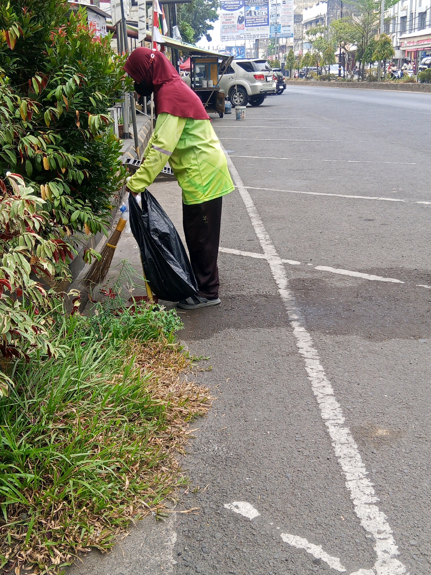 A woman wearing a maroon hijab and a lime green long-sleeved shirt is sweeping debris from a sidewalk into a black garbage bag. She is also holding a broom. The woman is standing next to a lush green bush with red-tipped leaves. In the background, a street with parked cars and a food cart is visible. The scene appears to be on a street in Lubuklinggau, Indonesia.