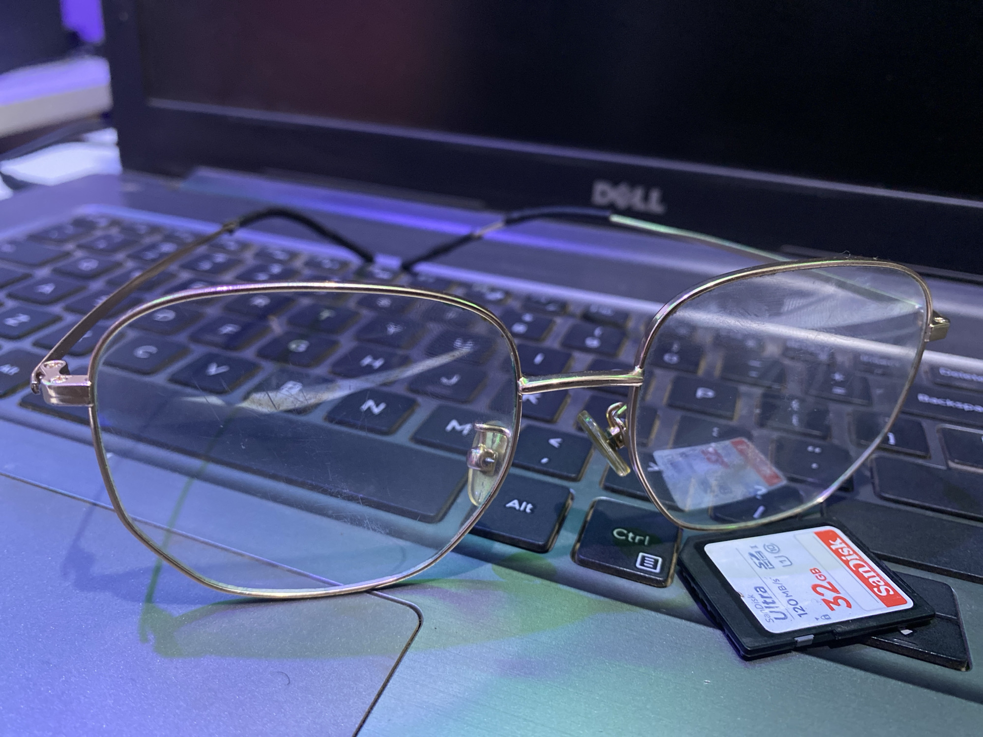 The image presents a close-up view of eyeglasses resting on a laptop keyboard, with a SanDisk 32GB SD card placed near the Ctrl key. The eyeglasses feature a gold-colored frame. The SD card displays the SanDisk logo, capacity, and speed specifications. The laptop is identifiable as a Dell product, indicated by the logo at the top right. The keyboard is black, and the overall lighting suggests an indoor environment. There is no evidence to suggest the location is in the city of Bukuru, Nigeria. There are no people in the image.