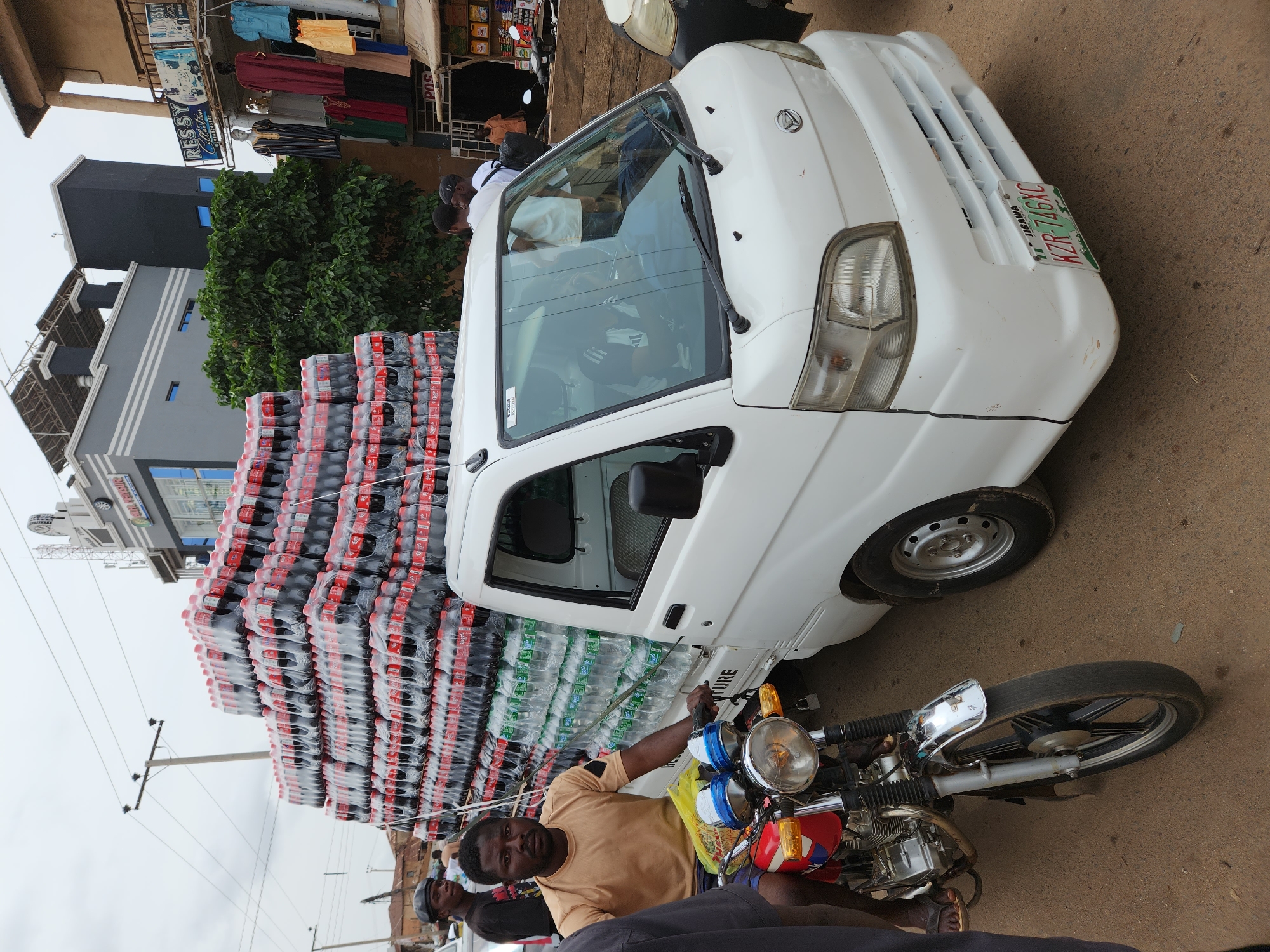 The image shows a white Daihatsu Hijet truck overloaded with stacks of Coca-Cola and bottled water in Jos, Nigeria. A man in a mustard-colored shirt is on a motorcycle parked near the truck. Other people are in the background. Buildings and businesses are visible in the background, suggesting an urban setting. The sky is overcast.