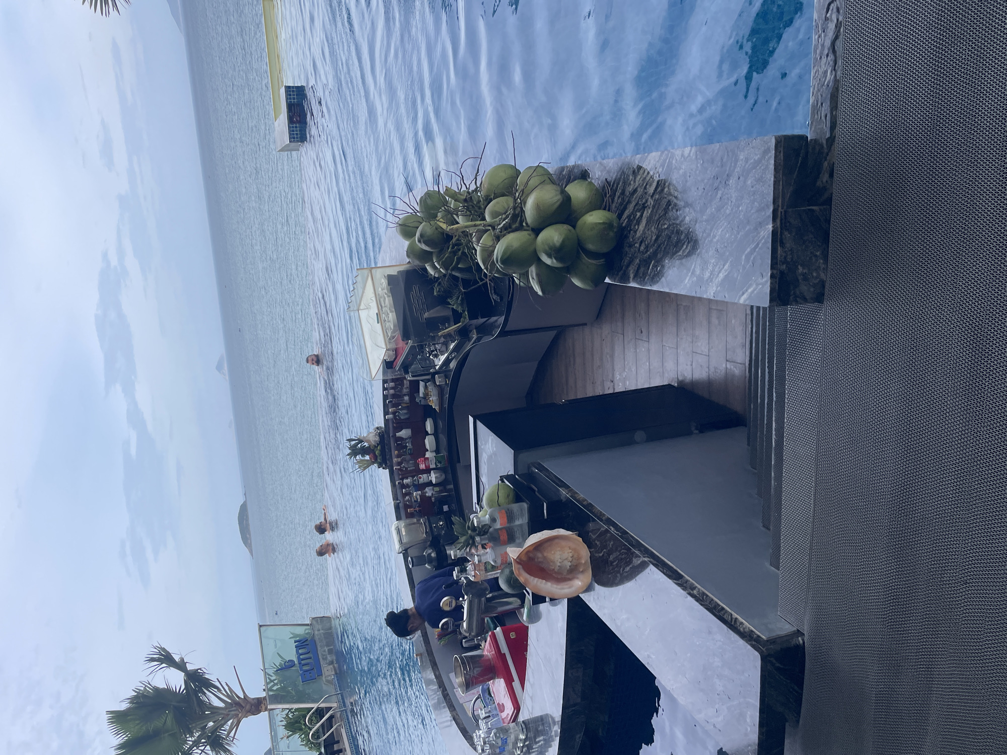 An exterior, eye-level, medium shot captures a scene at the Cotton Beach Club in Nha Trang, Vietnam. The image focuses on an infinity pool with the ocean in the background, under an overcast sky. In the foreground, a bar is set up with bottles of alcohol, a blender, and a tap. A bartender, wearing a dark blue shirt and with their hair in a bun, is visible behind the bar, attending to their work. A cluster of green coconuts and a large seashell are placed on the edge of the bar. Several people are seen swimming and relaxing in the pool, with a palm tree and a sign that reads "COTTON" visible on the left side of the frame. The pool's edge transitions to steps leading down to a textured gray matting.