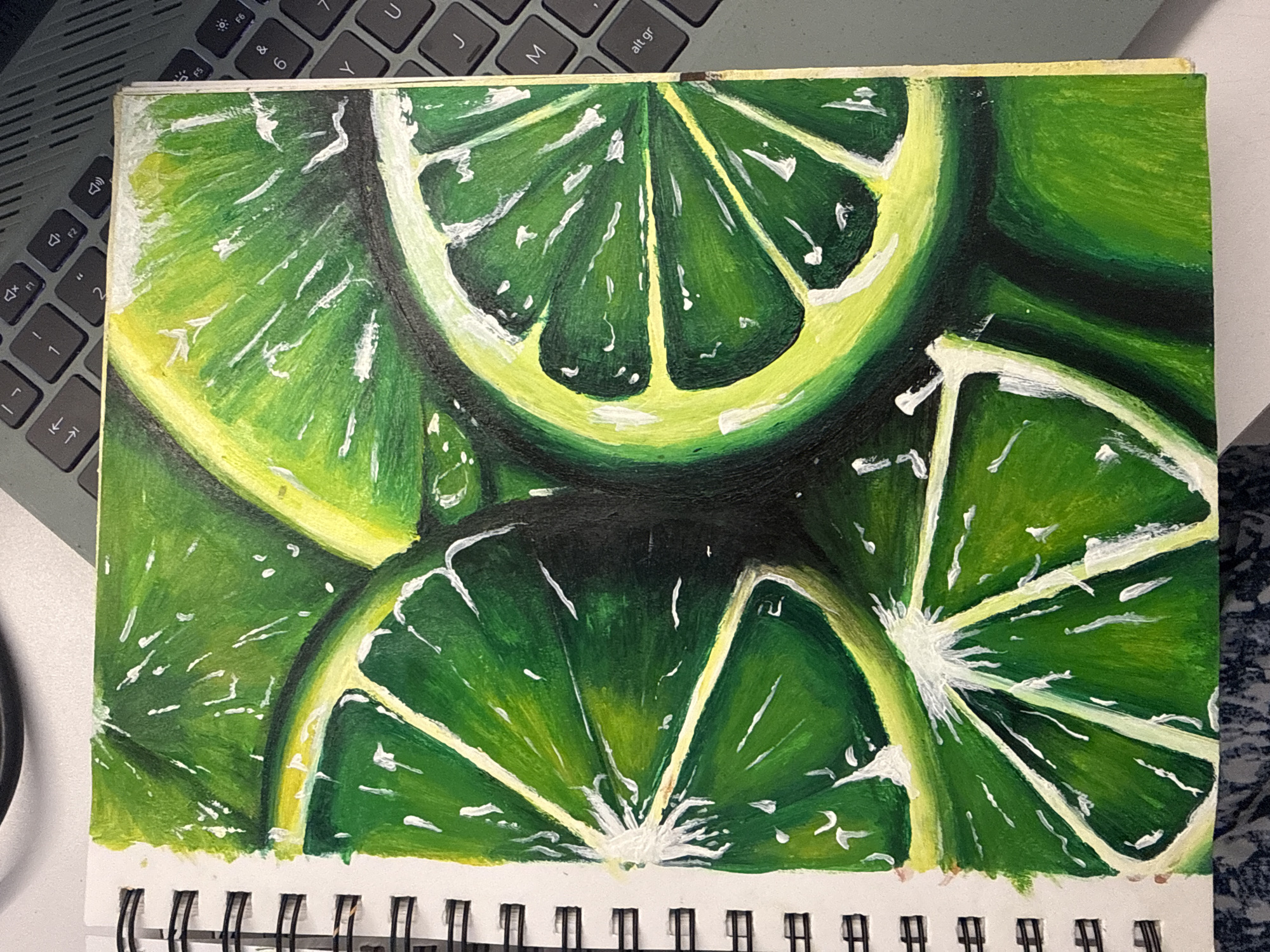 The image is a close-up, high-angle view of an open sketchbook filled with an original painting of lime slices. The painting depicts four lime slices in a semi-overlapping arrangement. The slices are rendered in shades of green and yellow, with white accents to highlight texture and juice. The sketchbook is lying on a dark grey laptop keyboard. The background is a light grey surface. The scene does not depict any location in Puebla, Mexico.