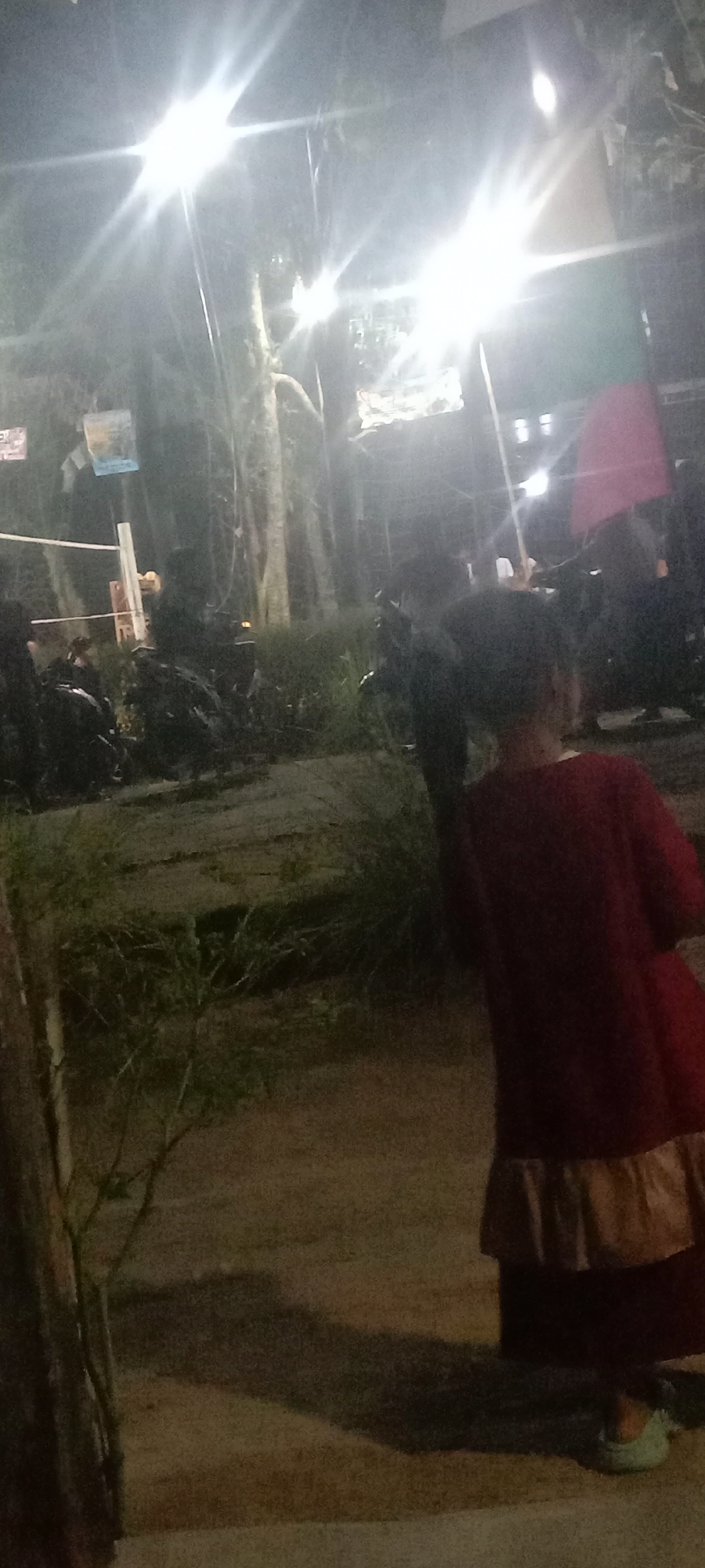 The image depicts a nighttime outdoor scene with bright artificial lighting. In the foreground, a person is visible from behind, wearing a red dress with a tan ruffled hem and light blue shoes. The person's back is to the camera, and they are facing away from the viewer and towards the left side of the frame. Behind this individual, there are dark shapes that appear to be motorcycles or scooters parked closely together, with some vegetation and trees visible in the background. The environment suggests an area with amenities for parking, possibly near a public space or event. Signs are visible in the upper left quadrant, partially obscured by light glare. The lighting creates strong specular highlights, particularly from overhead light sources which are not fully visible. The overall impression is a well-lit, possibly busy, outdoor area at night, with a focus on the individual in the foreground.