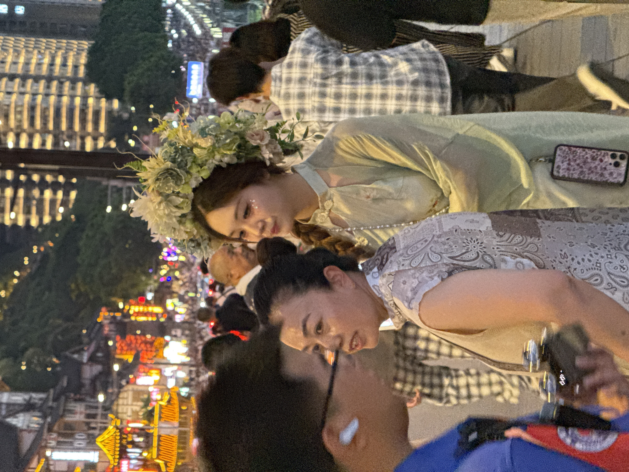 This image depicts an evening street scene in the Yuzhong District of China, likely during a festival or celebration, given the illuminated traditional Chinese architecture in the background and the festive attire of some individuals. In the foreground, a young woman wearing a large floral headdress and a light-colored traditional dress is the primary subject. She has glitter applied to her cheeks. To her left, a woman in a patterned blouse is visible, appearing to be in conversation with a man whose face is not fully visible, but who has dark, curly hair and is wearing glasses and an earbud. Behind these individuals, a crowd of people are seen walking, many dressed in casual attire, including one person in a grey and white plaid shirt. The overall atmosphere is lively, with numerous lights from buildings and street vendors creating a warm, vibrant ambiance. The Yuzhong District, known for its unique hilly topography and historic architecture, provides a distinctive urban backdrop to this scene.