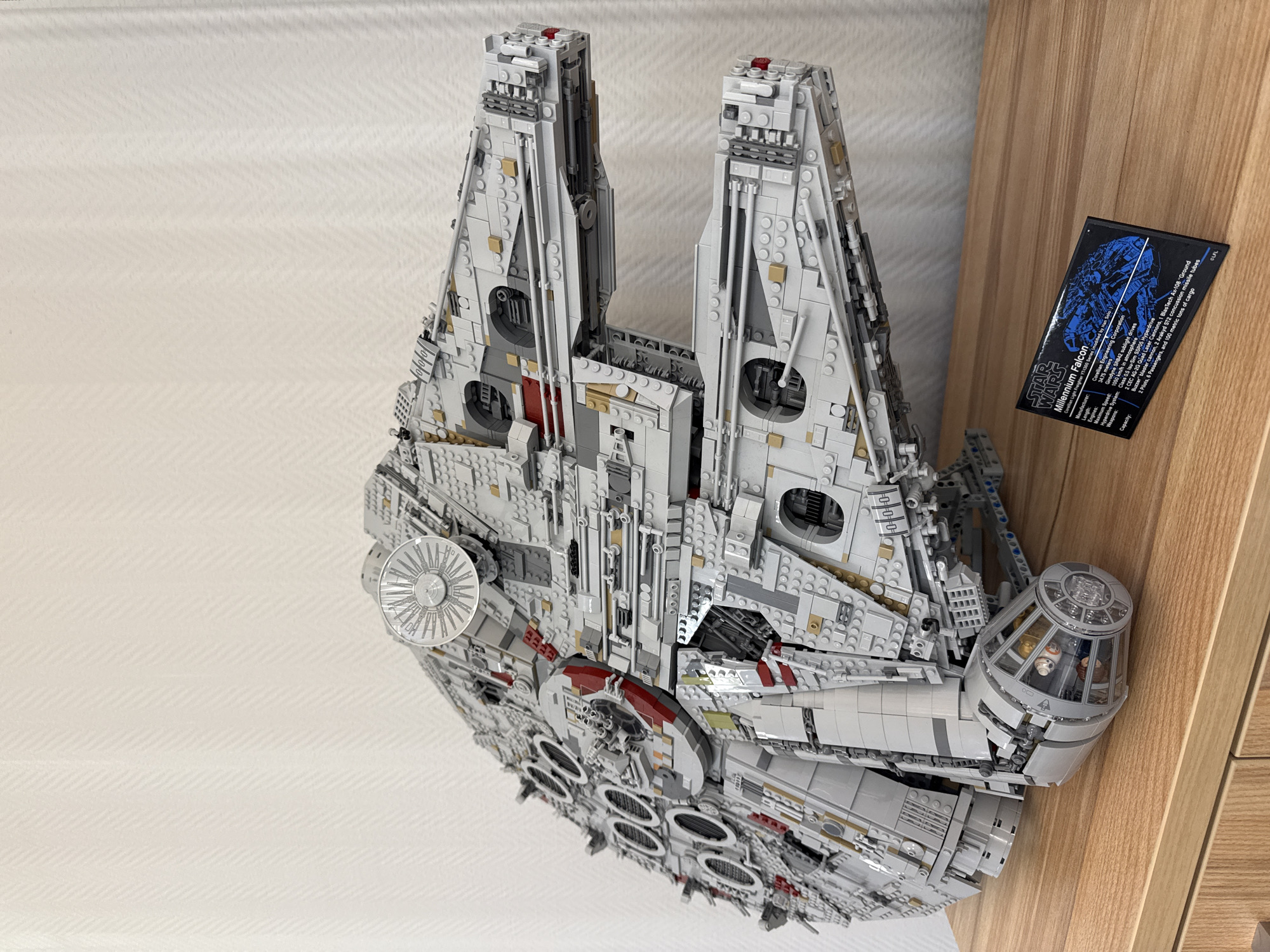 The image shows a Lego model of the Millennium Falcon from Star Wars. The model is large and detailed, resting on what appears to be a wooden surface next to a black rectangular card. The card features a blueprint-style image of the Millennium Falcon and text describing the ship's specifications, including its manufacturer, length, engine type, hyperdrive system, weapons, and capacity. The background is a white wall with a subtle texture. The scene is indoors and well-lit. There is no indication that this is in Krasnoyarsk, Russia.