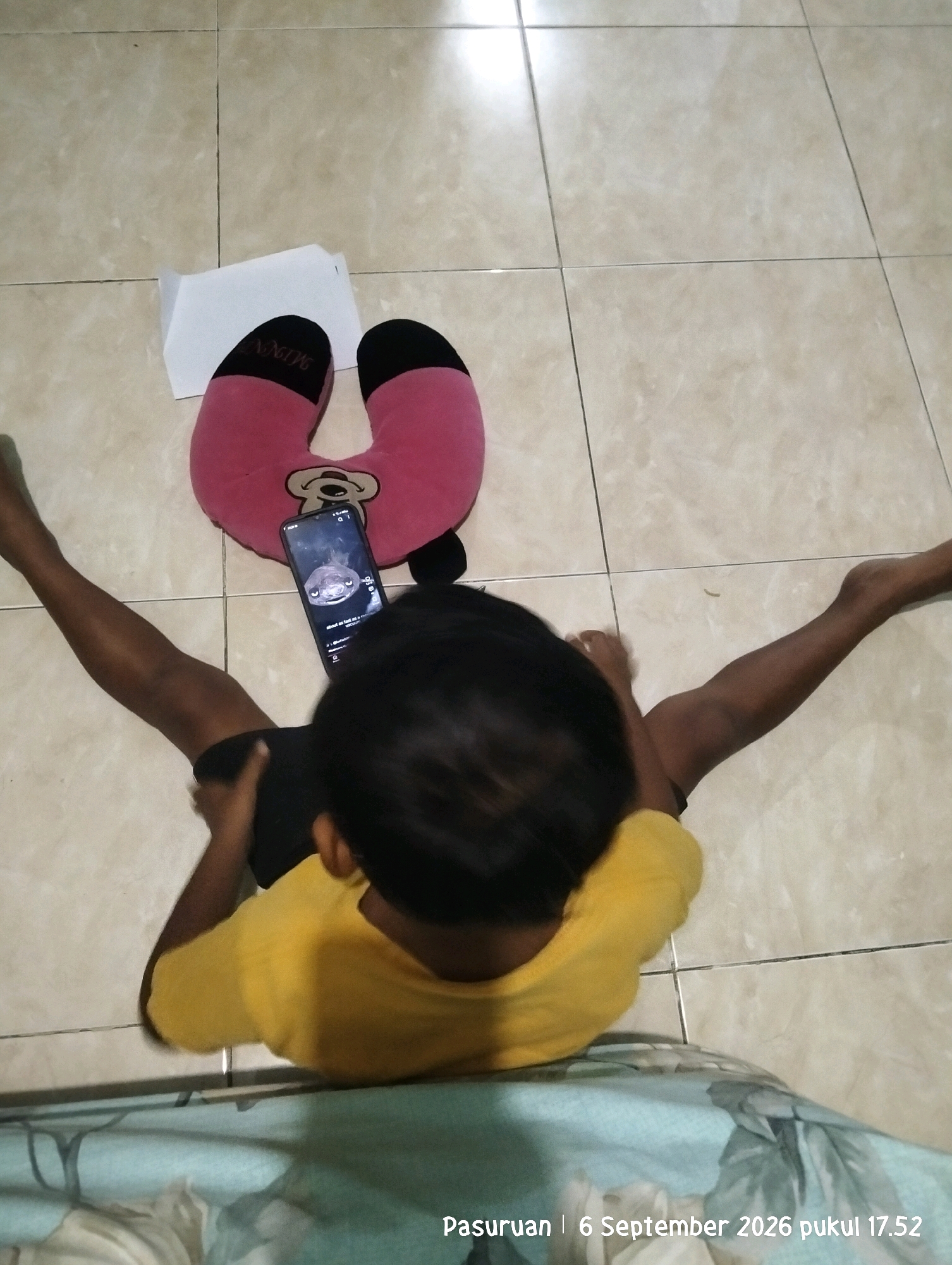 A young child wearing a yellow t-shirt lies on their back on a tiled floor, looking at a smartphone. The child's arms are outstretched, and their legs are bent. A pink U-shaped neck pillow with black accents and a Disney "Minnie" logo is positioned behind the child's head. A white paper is placed near the pillow. The floor is made of light-colored square tiles. A teal-colored patterned fabric is visible at the bottom of the frame. A timestamp in the lower right corner reads "Pasuruan | 6 September 2026 pukul 17.52".