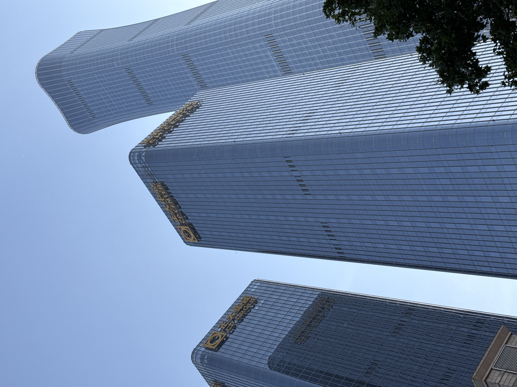The image depicts a low-angle view of three modern skyscrapers in the Chaoyang District of China. The buildings have a smooth, curved design and are primarily constructed of glass and metal. The sky is light blue and mostly clear. The top of a tree is visible on the right side of the image. The logos of the "CR CENTER" are visible on two of the buildings.