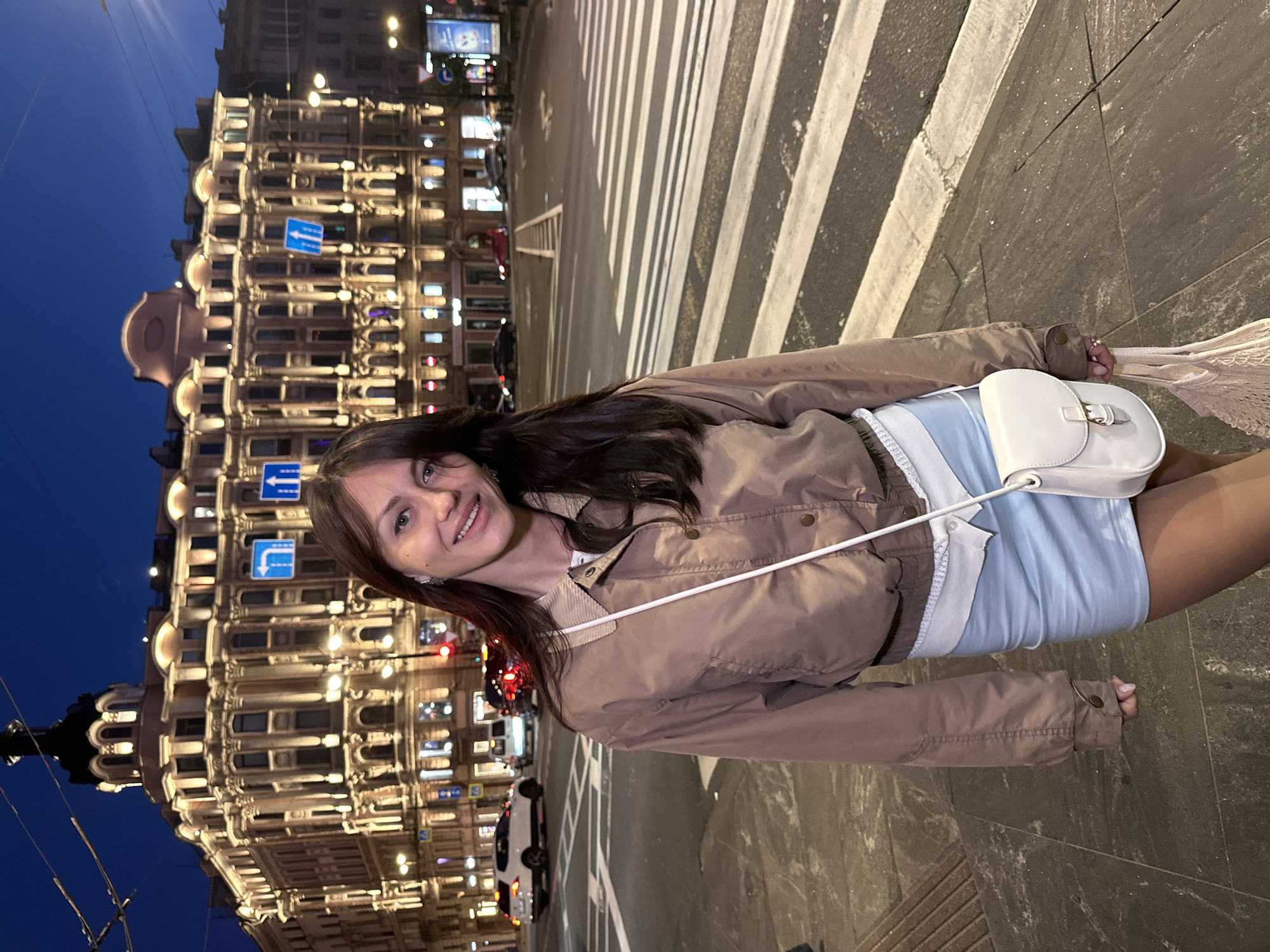A young woman stands on a crosswalk in Saint Petersburg, Russia, at dusk. She has long, dark hair and is wearing a brown jacket, a light blue skirt, and a white purse. Behind her, a large, ornate building is illuminated by warm lights, with traffic signs and streetlights visible in the background. Cars are driving on the street, and the sky is a deep blue. The scene appears to be a busy urban area.