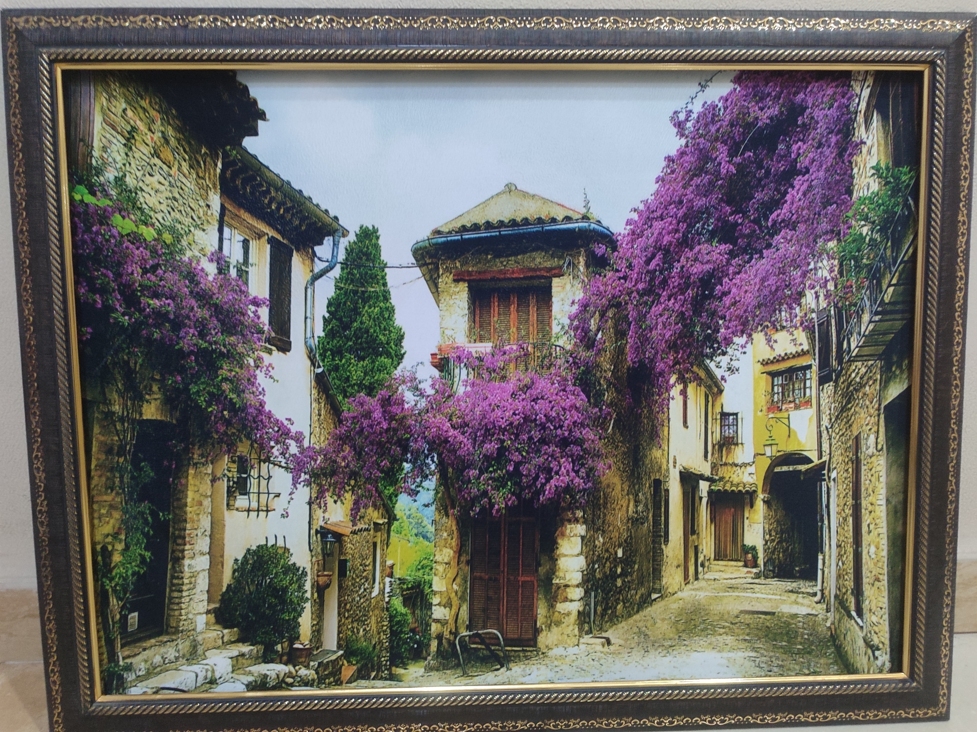This is a framed image of a narrow, cobblestone street in what appears to be a European village. The buildings are old and made of stone, with terracotta tile roofs and wooden shutters on the windows. Vibrant purple bougainvillea vines are cascading down the sides of the buildings, adding a splash of color to the scene.
The street is paved with cobblestones, and there are a few stone steps leading up to doorways. A small, dark-colored bench is placed in front of a wooden door. The overall atmosphere is quaint and picturesque, suggesting a peaceful and idyllic setting. The time of day appears to be daytime, with bright sunlight casting shadows on the street. There are no people visible in the image.
The frame is dark brown with a decorative gold trim. The image depicts a charming and picturesque scene that evokes a sense of old-world charm and tranquility. No text is visible in the image or the frame.