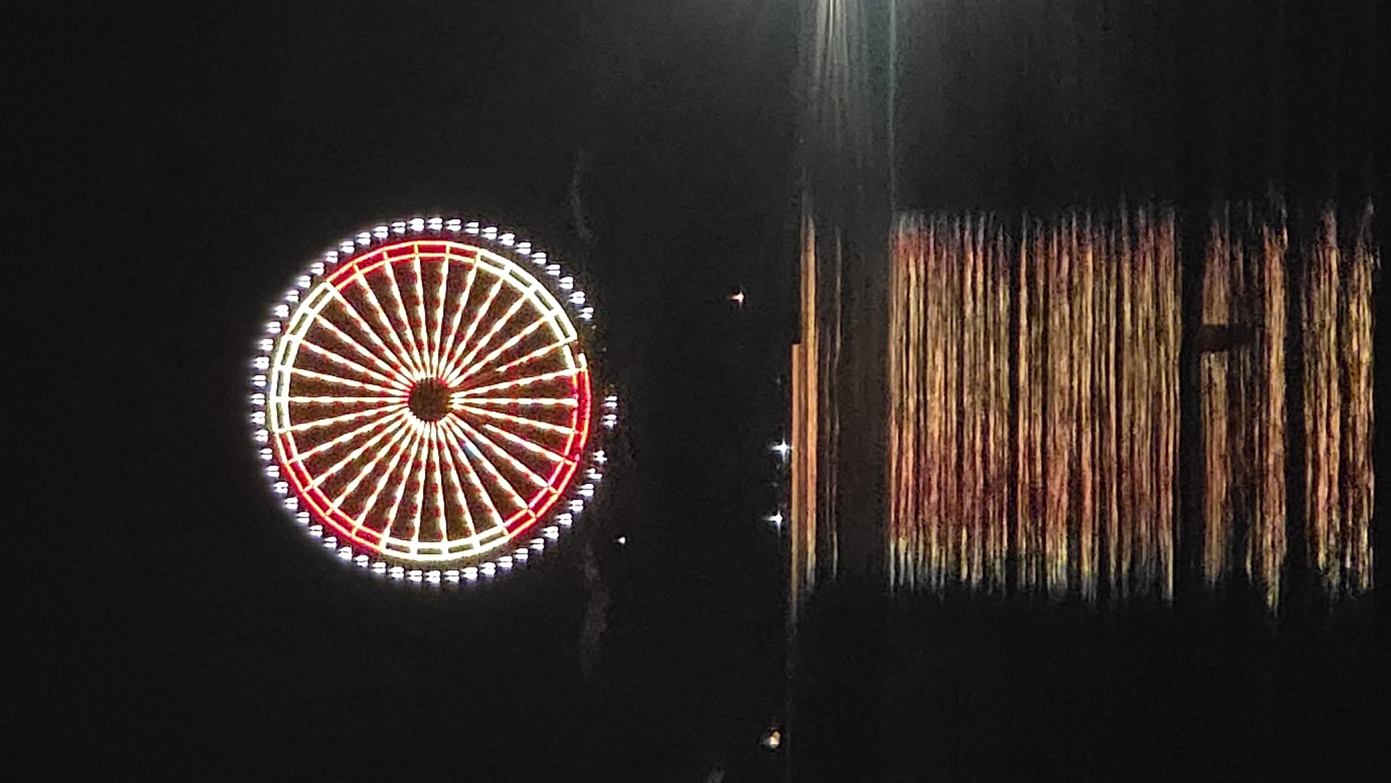 The image shows a lit Ferris wheel and its reflection in water, taken at night. The Ferris wheel has a white outer rim with lights and red spokes. The reflection consists of distorted vertical lines of red, yellow, and orange. The background is dark, indicating it is nighttime. The location is likely Nha Trang Ward, Vietnam.