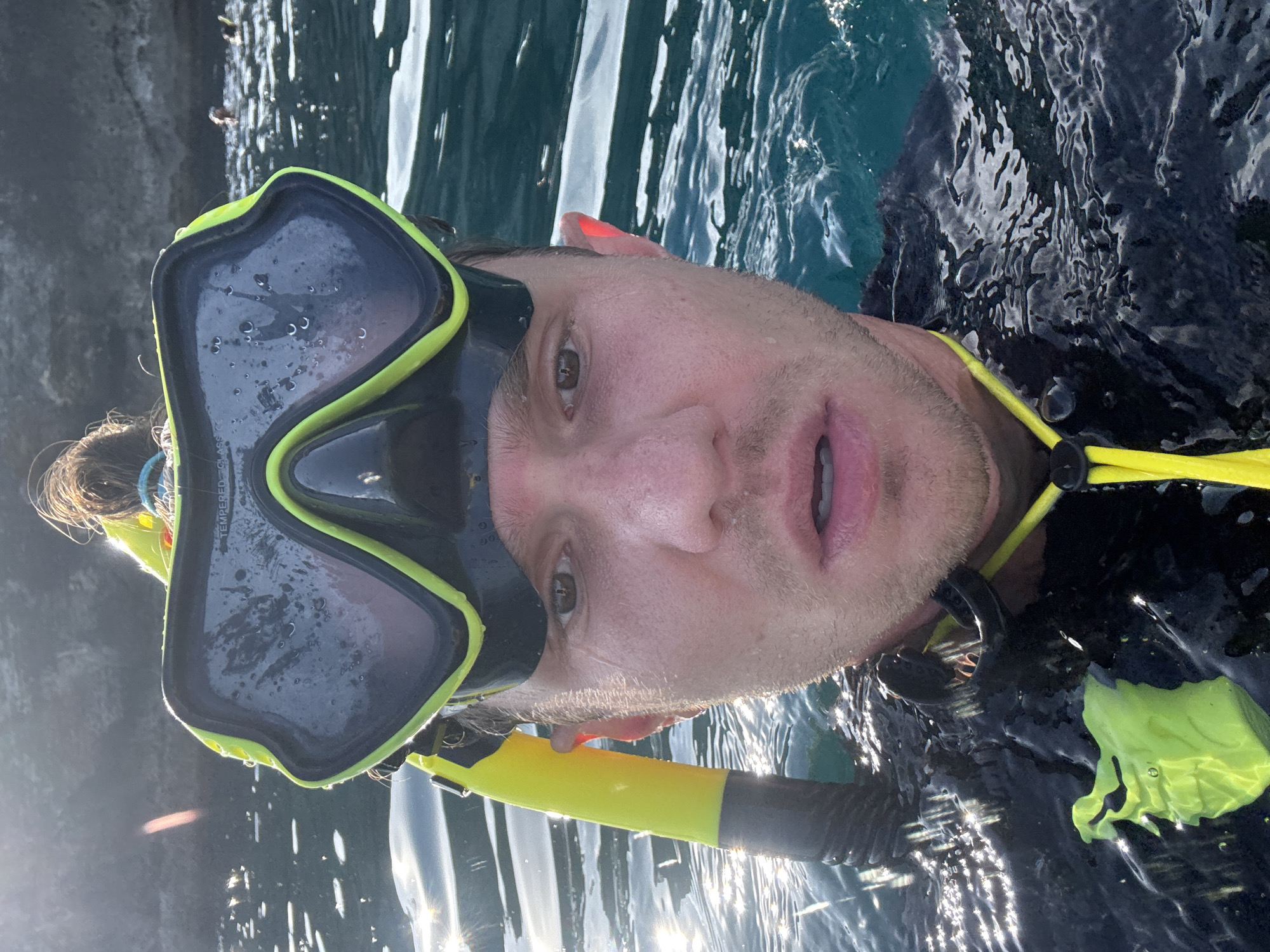 The image shows a person in the water with snorkeling gear, including a mask with a yellow and black frame, a snorkel, and a wetsuit. The person is looking directly at the camera. The water is reflecting sunlight, and there is a rocky cliff face visible in the background. Although I cannot pinpoint the specific location in Nusa Penida without additional landmarks, the image suggests a coastal area suitable for snorkeling.