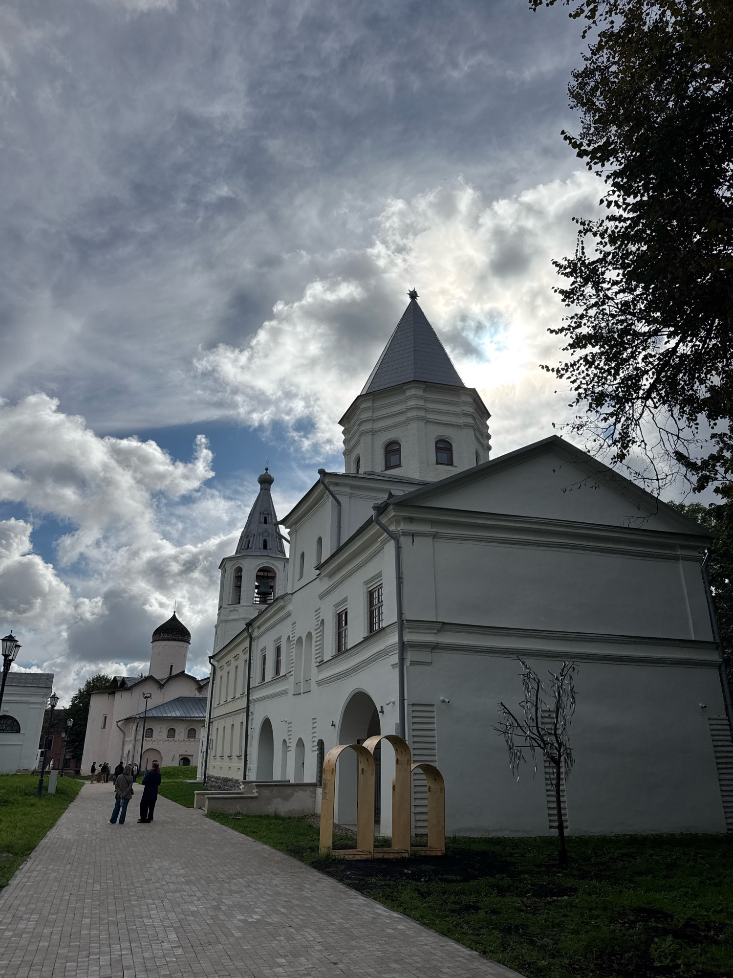 The image depicts the Veliky Novgorod Kremlin, also known as Detinets, in Veliky Novgorod, Russia. The scene captures a path paved with light-colored bricks leading towards several white architectural structures. In the foreground, two individuals, one wearing a dark jacket and the other a grey hoodie, are walking away from the camera on the path.
To the left of the path, a lamppost stands, and further back, other buildings and a darker-domed structure are visible. The main focus of the image is on the white buildings with red-trimmed windows, featuring arched entrances and tiered roofs. One prominent tower, characteristic of Russian Orthodox churches, has a grey conical roof topped with a small sphere and a star. Another, taller tower, also grey and conical, has arched openings where a bell is visible. The architecture suggests a historical and religious complex.
In front of the white building on the right, a small, bare tree adorned with reflective strips hangs from its branches. Beside the tree, two golden, arch-like structures stand on the grass. The ground around the buildings is covered with green grass, and the sky above is dramatic, with a mix of dark and bright clouds, and the sun partially visible, casting rays through the overcast. A large tree with dark foliage partially frames the upper right corner of the image.