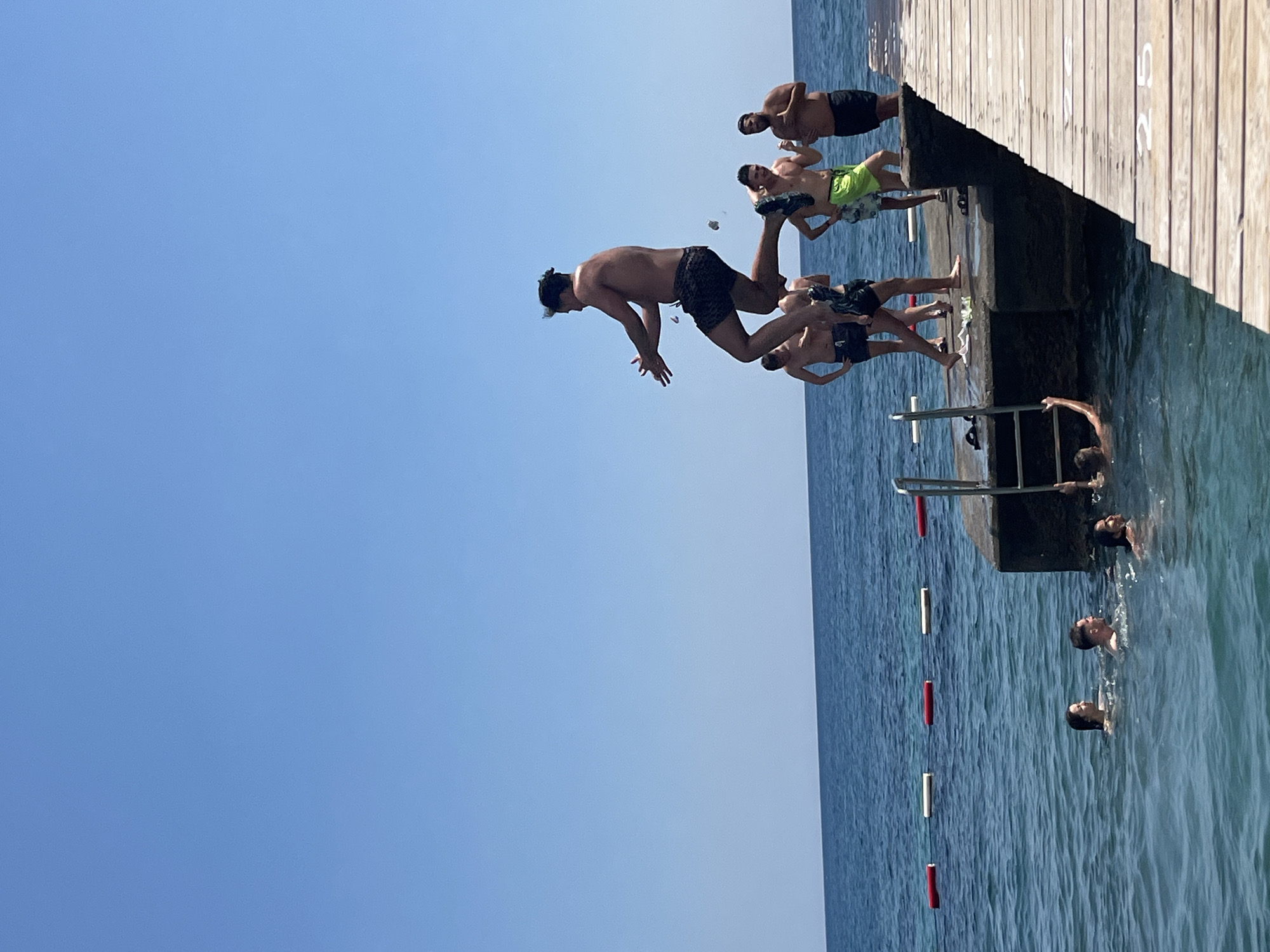 At a beachside pier in Budva, Montenegro, a group of young men are engaged in recreational activities. The sky is a clear, bright blue, and the sea is a deep blue. A wooden pier extends into the water, with a concrete section at the end. Several men are on the pier, with one in mid-air performing a jump into the water. Others are standing, watching, or preparing to jump. In the water, several people are swimming and enjoying the sea. Buoys mark a swimming area in the sea.