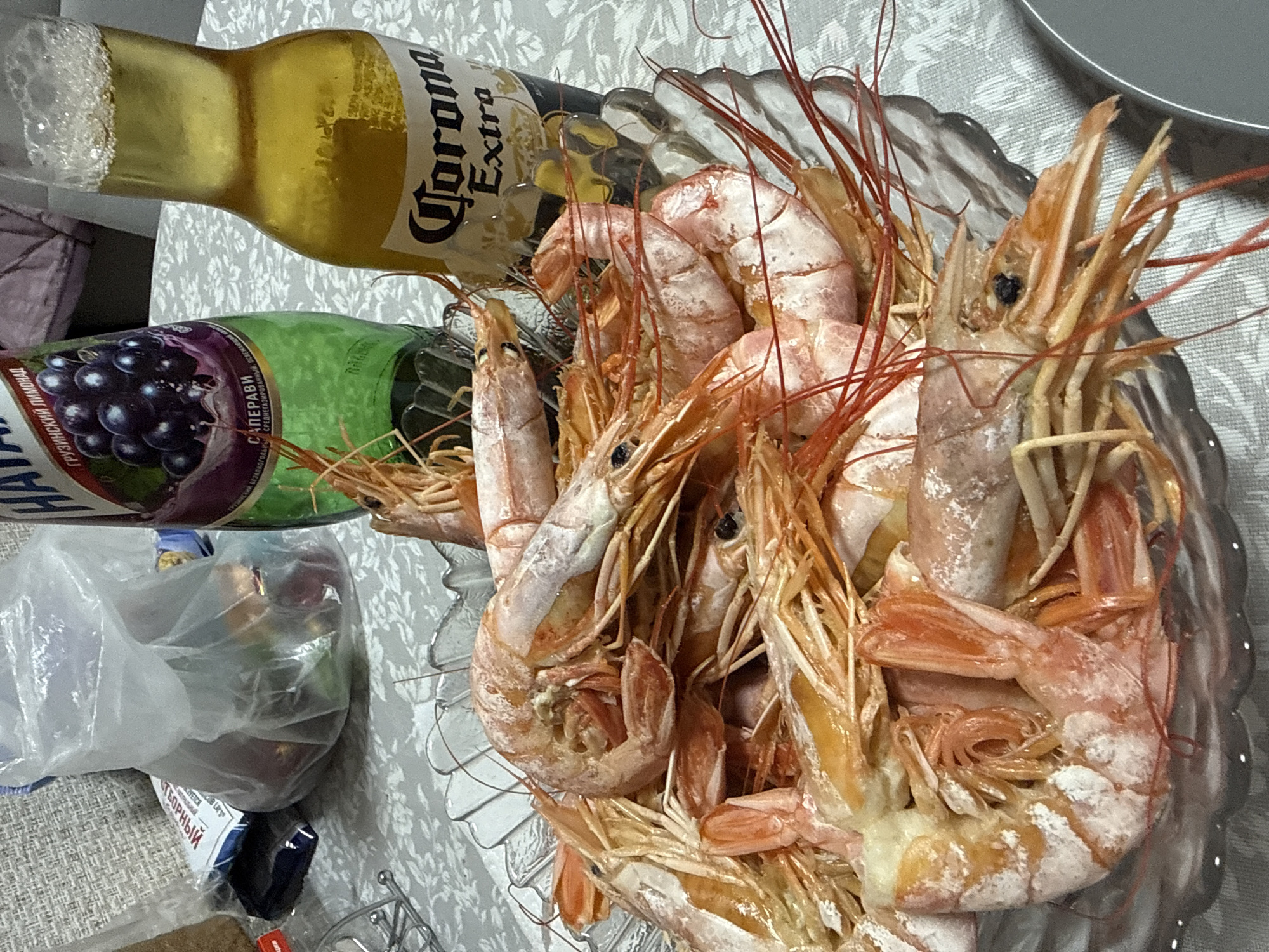 The image shows a bowl of cooked shrimp, a bottle of Corona Extra beer, and a bottle of grape juice on a table covered with a patterned tablecloth. The scene is indoors, likely in a private residence. Details suggest a casual meal setting.