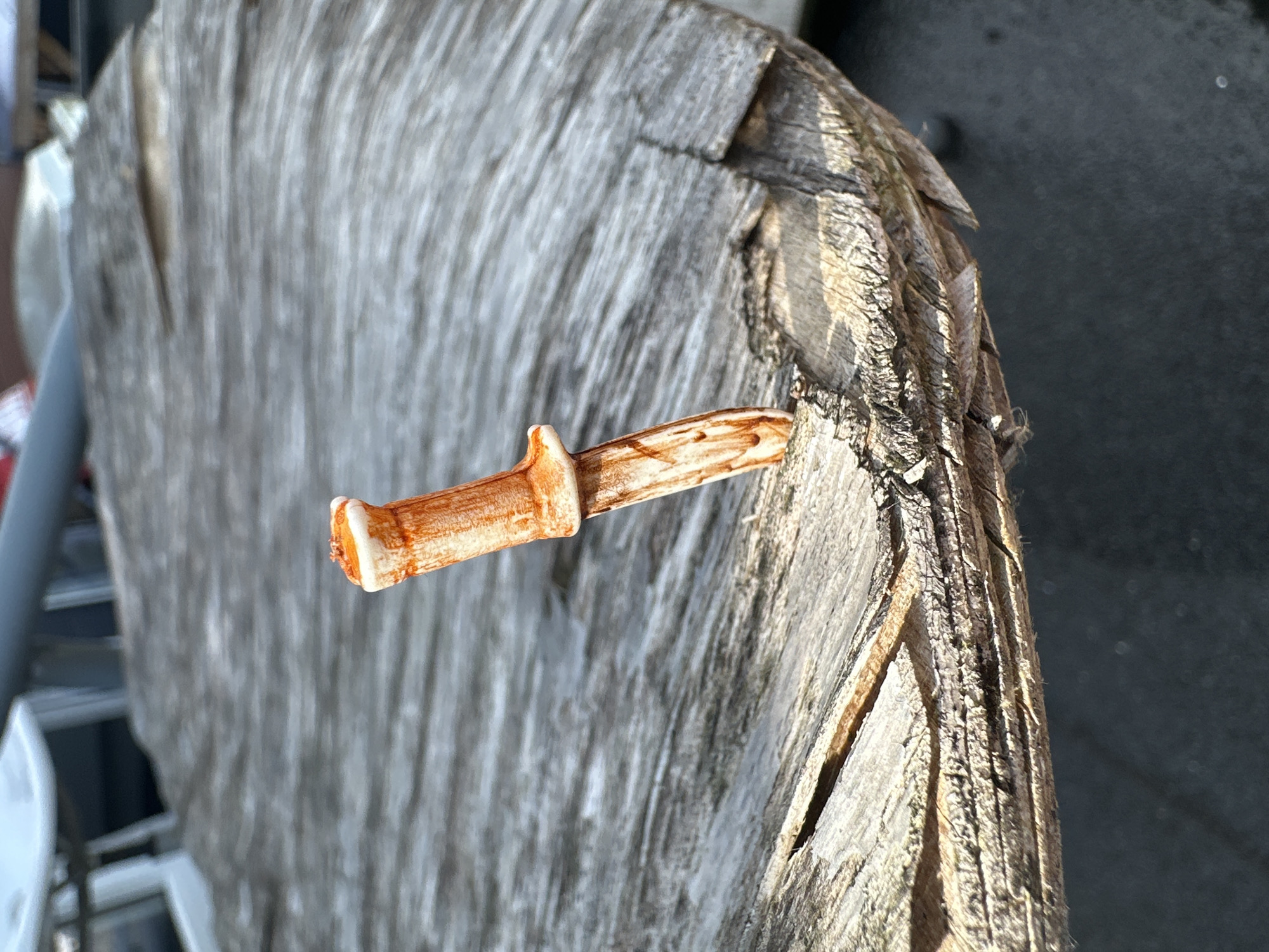 A miniature ceramic knife, with an orange and white handle, is embedded in a weathered, gray wooden surface. The knife appears to be stuck at an angle, with its blade extending into the wood. The wooden surface has a rough texture with visible grain and some cracks. The background is out of focus, showing hints of outdoor furniture and a dark, paved area. The lighting suggests it is daytime, with sunlight casting shadows and highlighting the textures of the wood. The scene does not provide any information about a specific location in Lelystad, Netherlands.