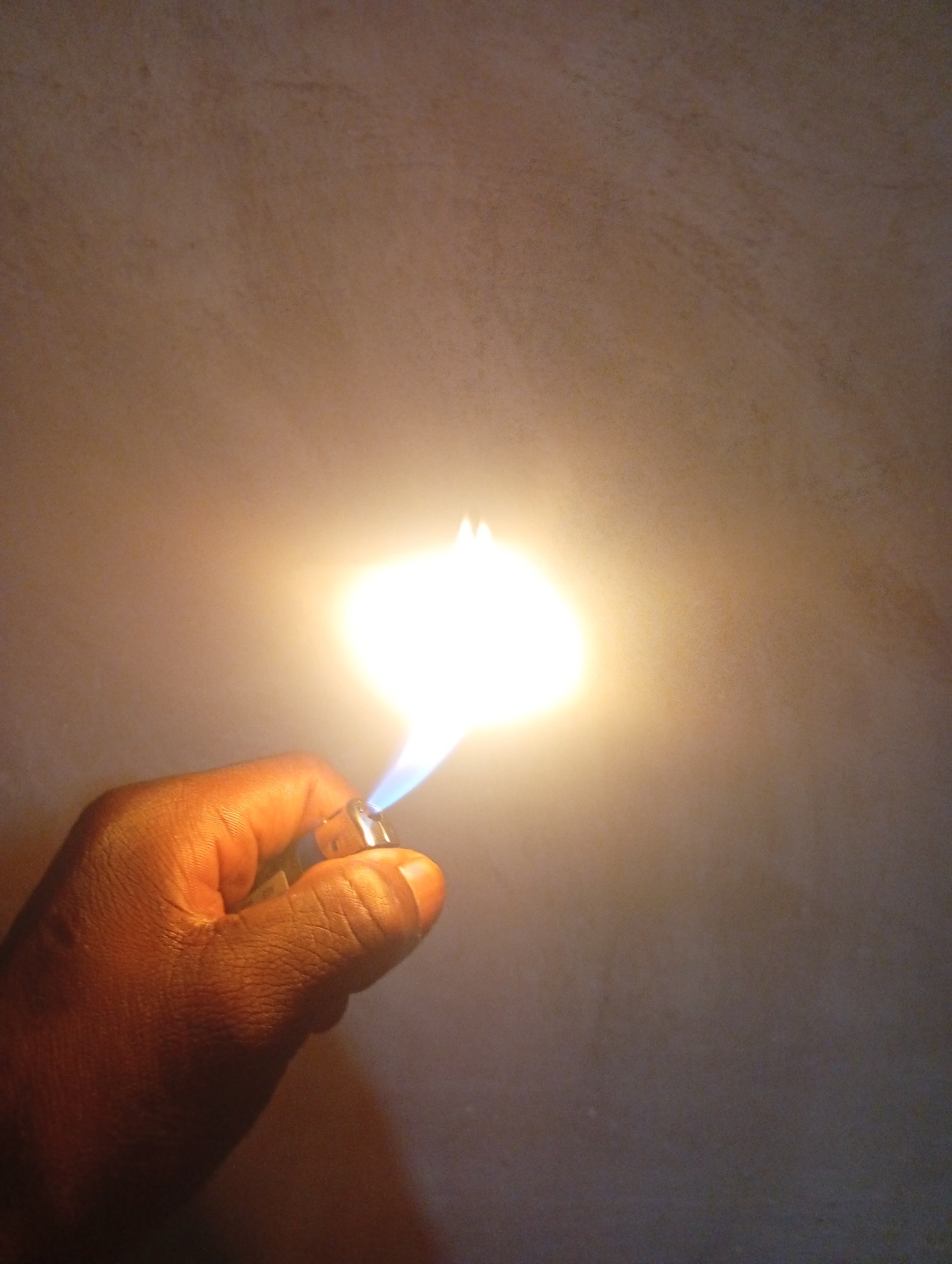 A close-up shot shows a dark-skinned hand holding a metal lighter, which is emitting a bright flame. The flame is predominantly white and yellow, with a blue base near the nozzle of the lighter. The background is a plain, textured wall that is illuminated by the light from the flame, creating a gradient of light and shadow. The overall lighting of the image is dim, with the flame serving as the primary light source. The image does not provide any identifiable landmarks or context to determine a specific location within a city.