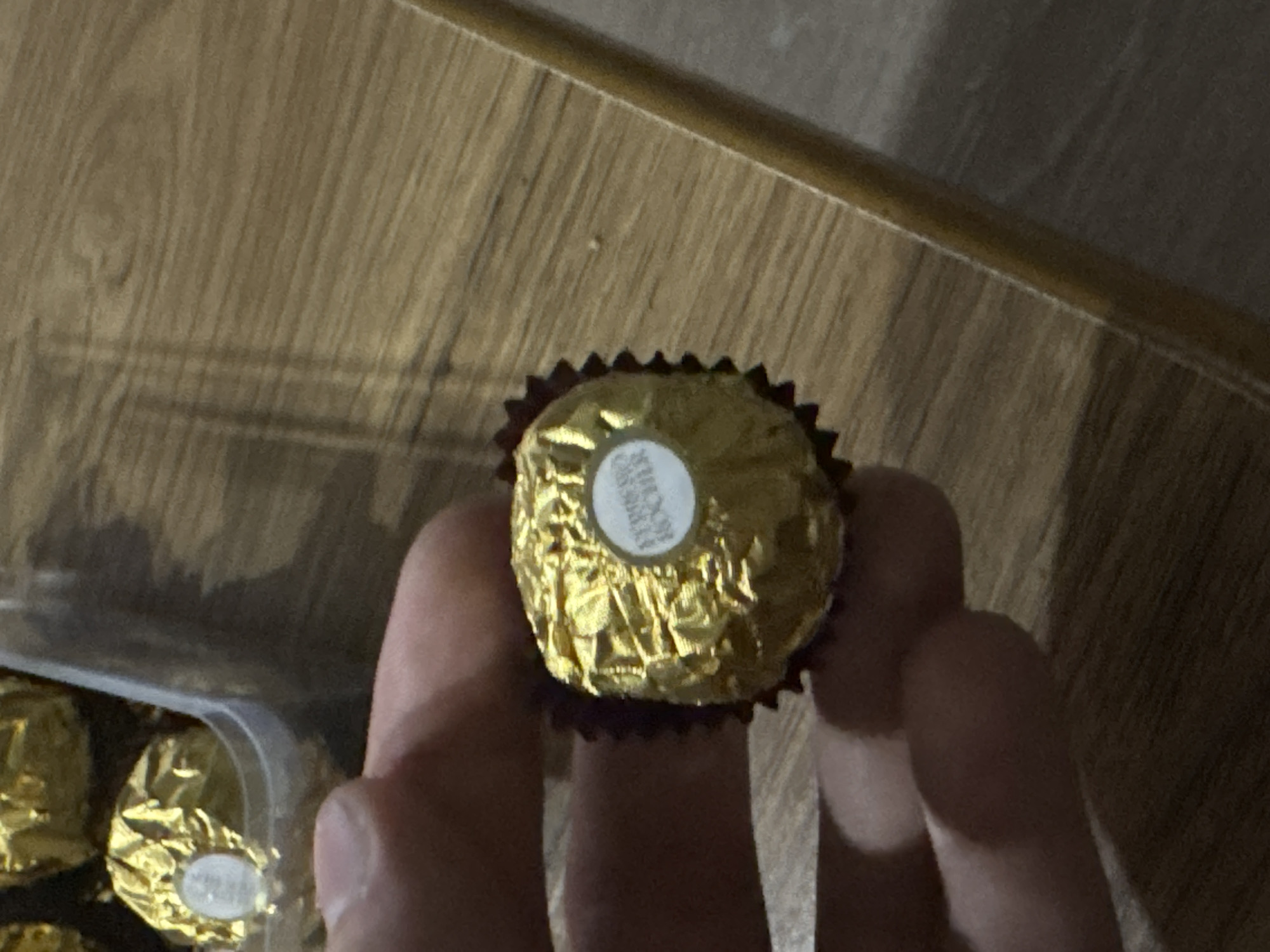A close-up, eye-level shot captures a hand holding a Ferrero Rocher chocolate. The chocolate is wrapped in its signature gold foil, with a white circular label clearly visible on its surface, bearing the brand name "Ferrero Rocher" in dark text. The hand's fingers are gently cupping the chocolate from below, obscuring a portion of the foil. The background features a wooden surface with a visible grain pattern, suggesting a table or countertop. In the upper left corner, a portion of a clear plastic container holding more Ferrero Rocher chocolates is visible, indicating that the chocolate being held is part of a larger assortment. The lighting in the image creates shadows that fall across the wooden surface, hinting at an indoor setting. The scene is devoid of any specific landmarks or indicators that would place it within the city of Tbilisi, Georgia.