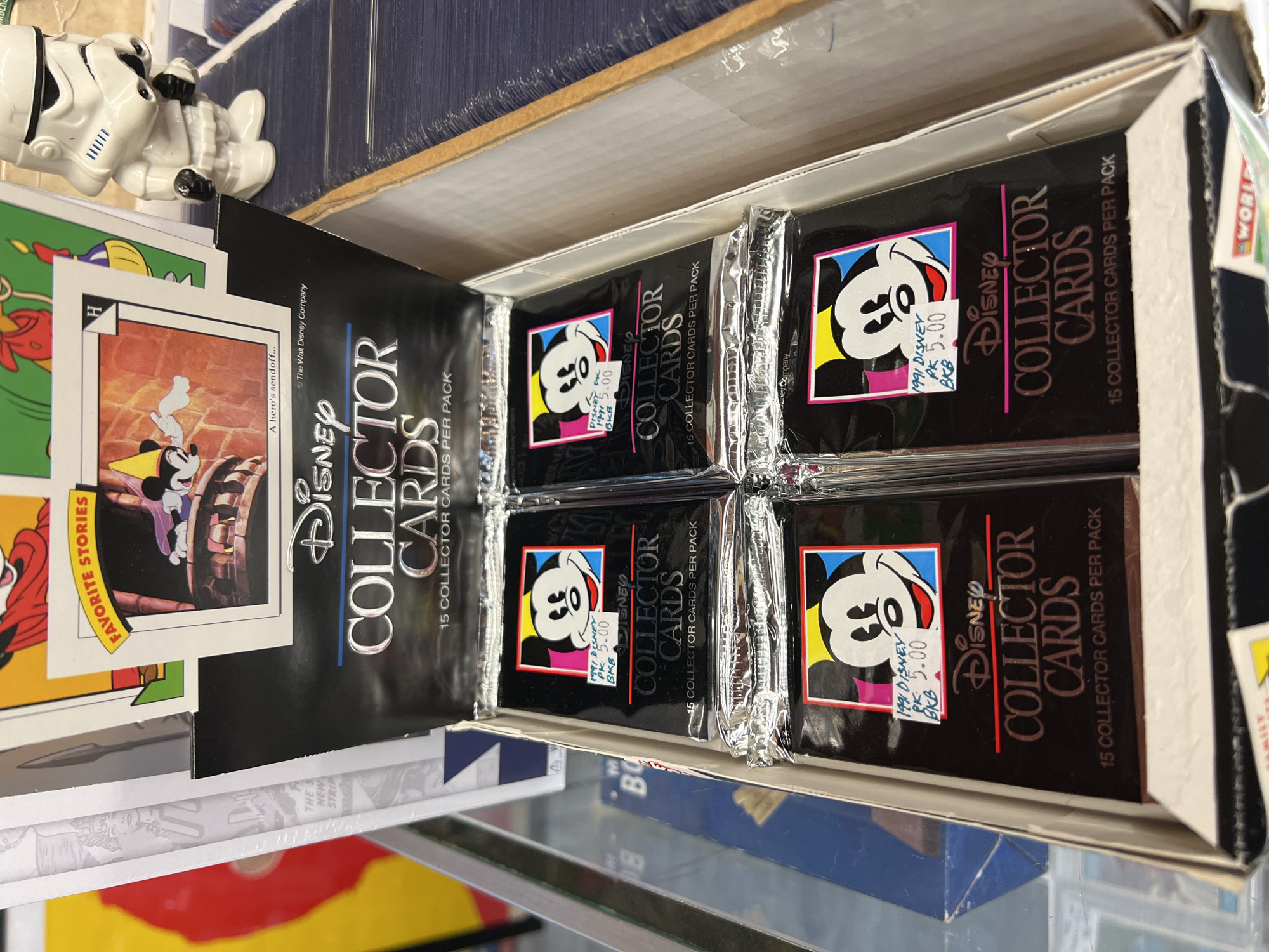 This image shows a box of unopened 1991 Disney Collector Cards in a store, likely a vintage or collectible shop, given the presence of other cards and items like the Stormtrooper figurine. The card packs feature Mickey Mouse on the front and are priced at $5.00 each. The location is identified as the City of Newburgh, United States of America, inferred from similar images available.