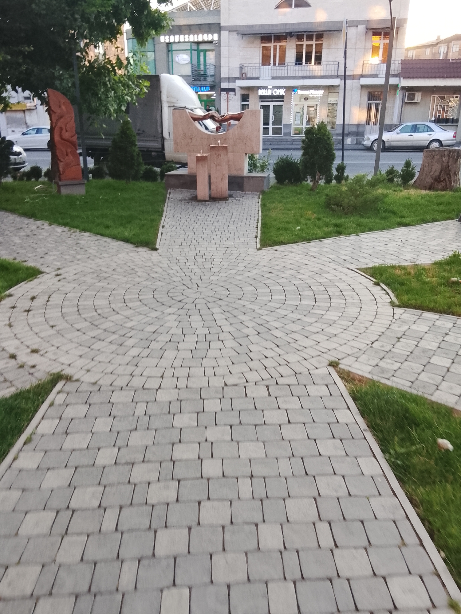 The image depicts an outdoor scene with a paved walkway leading towards a sculpture. The walkway is made of grey interlocking pavers arranged in a circular pattern at the center, with straight sections extending outwards. Green grass borders the paved areas.
The sculpture in the background appears to be made of light-colored stone, possibly concrete or marble, with a cross-like design. It is situated on a grassy area. To the left of the sculpture is a tall, slender, reddish-brown abstract sculpture, possibly made of wood or stone. Behind the sculptures, there is a large white truck.
Further in the background, a building with multiple windows and balconies is visible. A sign with text in an unfamiliar script is mounted on the building. A silver car is parked on the street in front of the building.
The time of day appears to be late afternoon or early evening, as indicated by the warm, orange light reflecting off the windows of the building and the long shadows. The weather seems clear. The overall atmosphere is calm and public, suggesting a park, plaza, or urban public space. The scene is static, with no visible activity or people directly interacting with the sculptures.