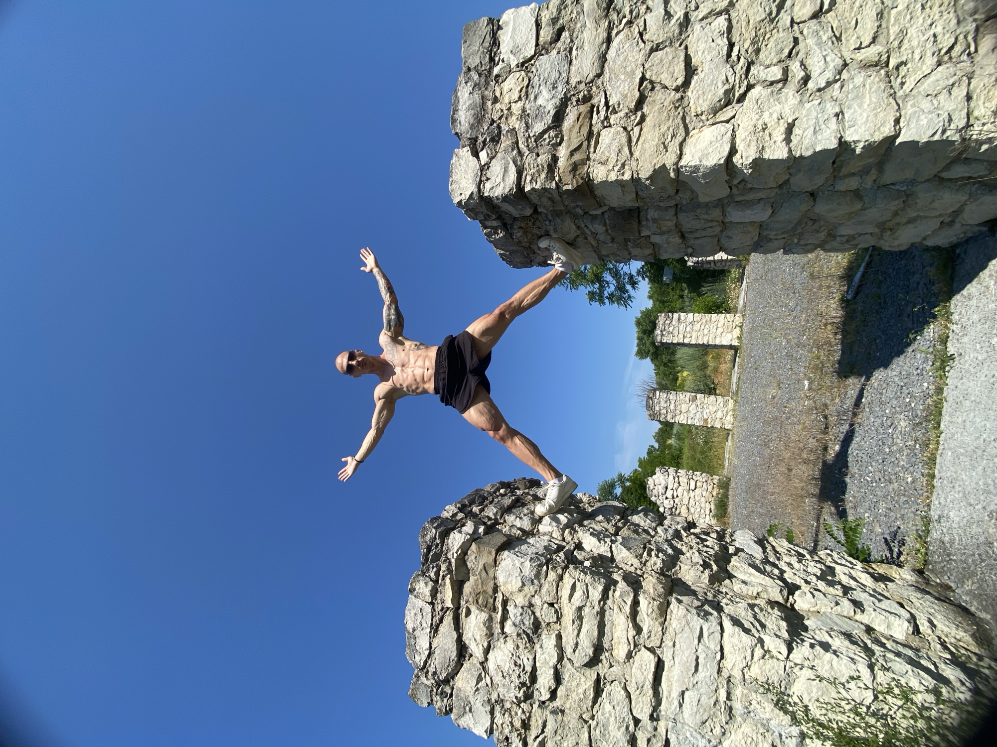 A shirtless man with a shaved head, sunglasses, and tattoos on his arms is captured mid-air, performing a wide split jump between two large stone ruins. He is wearing black shorts and white sneakers. His arms are outstretched to the sides. The ruins appear to be made of rough, weathered stones and are positioned in the foreground, framing the man. In the background, more stone structures and greenery are visible under a clear, bright blue sky. The ground is covered with gravel and dry grass. The scene is outdoors during the daytime. The location is the historical site of Hisarlaka Fortress in Pleven, Bulgaria.