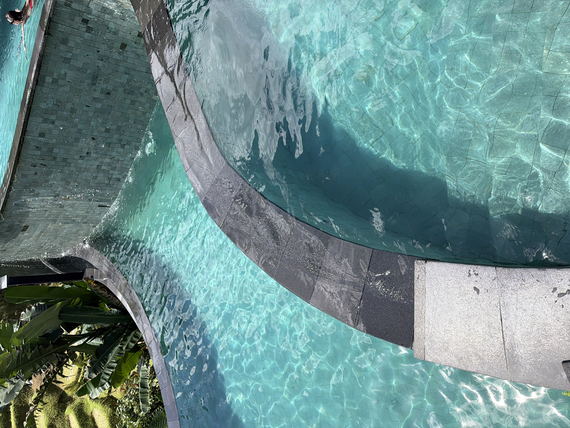 The image depicts a swimming pool with clear, turquoise water located in Tegal Suci, Indonesia. The pool has a tiled bottom and a dark gray tile border. A second pool, also with the same turquoise water, is connected by a cascade. Green tropical plants are visible near one section of the pool. A swimmer is partially visible in the top left corner of the image. Sunlight reflects off the water, creating light patterns on the pool floor.