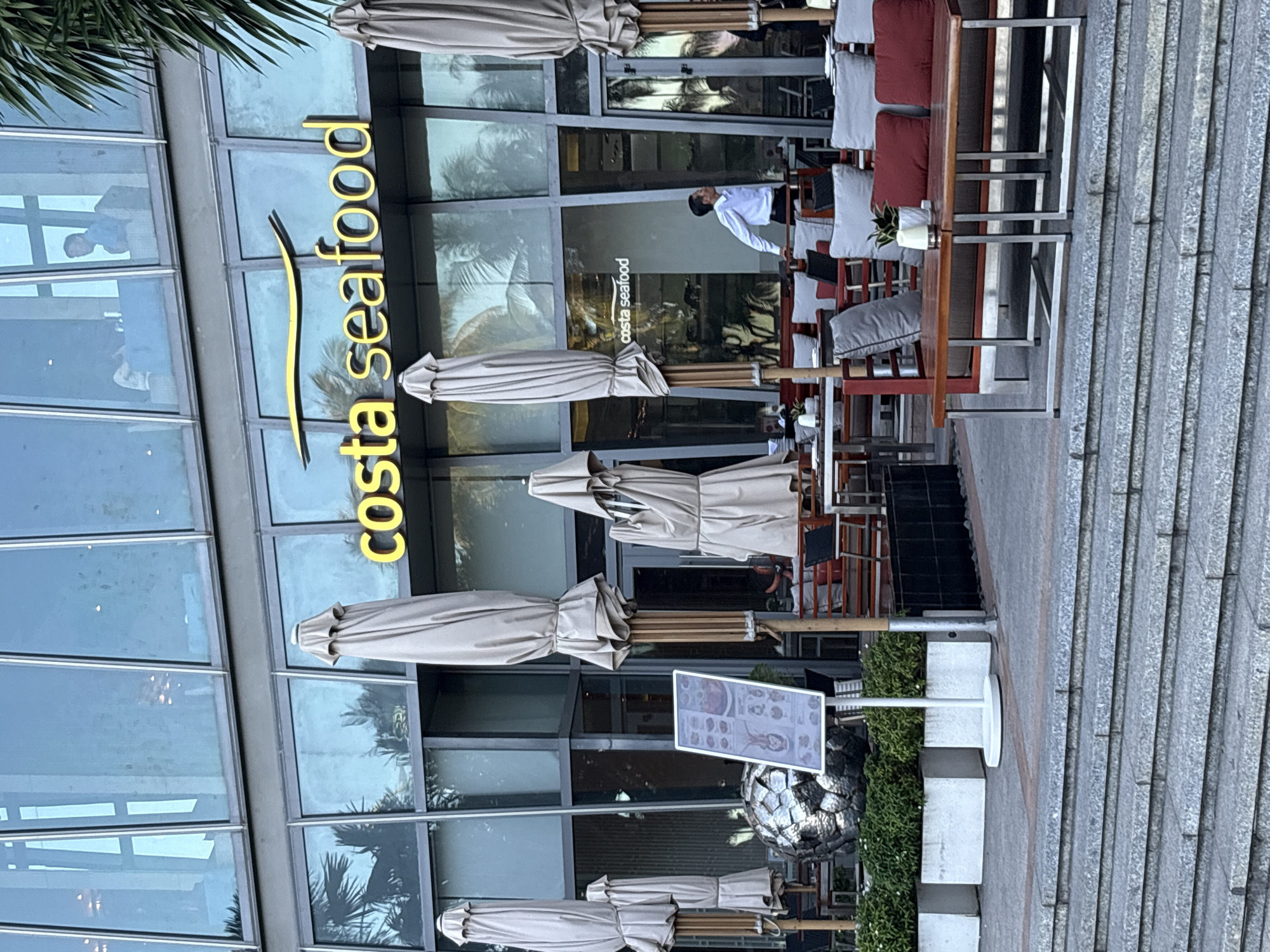 The image captures the exterior of a restaurant in Nha Trang Ward, Vietnam, with a prominent "costa seafood" sign in yellow lettering. The restaurant features large glass windows that reflect the surrounding environment, including palm trees and the sky. Outside, several closed, light-colored umbrellas are positioned, along with outdoor seating areas. The seating consists of tables and chairs, some with red and gray cushions. In the foreground, there are stone stairs leading up to the restaurant's entrance. A man in a white shirt and dark pants is visible through the glass, standing near a table. A menu stand is placed near the entrance. The overall scene suggests a daytime setting, possibly before opening hours or during a less busy period.