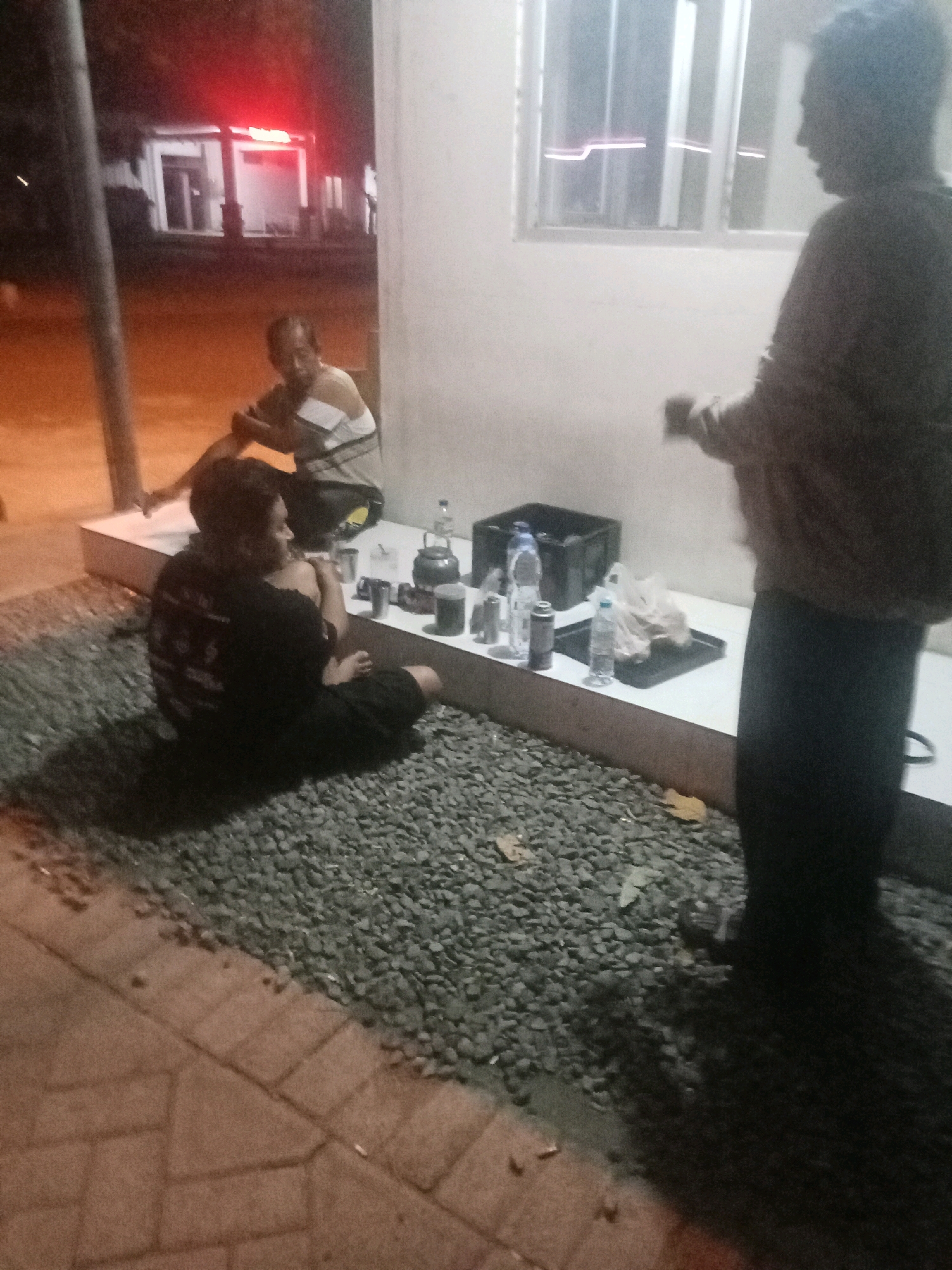At night, on a gravel-covered area in front of a building in Jombang, Indonesia, three people are present. The building has white walls and a window with horizontal white blinds. A light is visible within the building, possibly from a sign.
In the foreground, a person is sitting cross-legged on the gravel, wearing a black shirt and dark pants. To their left, another person is sitting on a raised white platform, wearing a striped shirt with white and dark bands. This person appears to be holding something.
Further to the right, a third person stands, facing away from the camera. They are wearing a light-colored, loose-fitting garment over dark pants.
On the white platform, several objects are arranged. These include bottles of water, cans, a dark plastic bin, a black kettle, and a plastic bag. The area is illuminated by artificial light sources, creating shadows on the ground. The ground in front of the gravel area is paved with interlocking bricks.