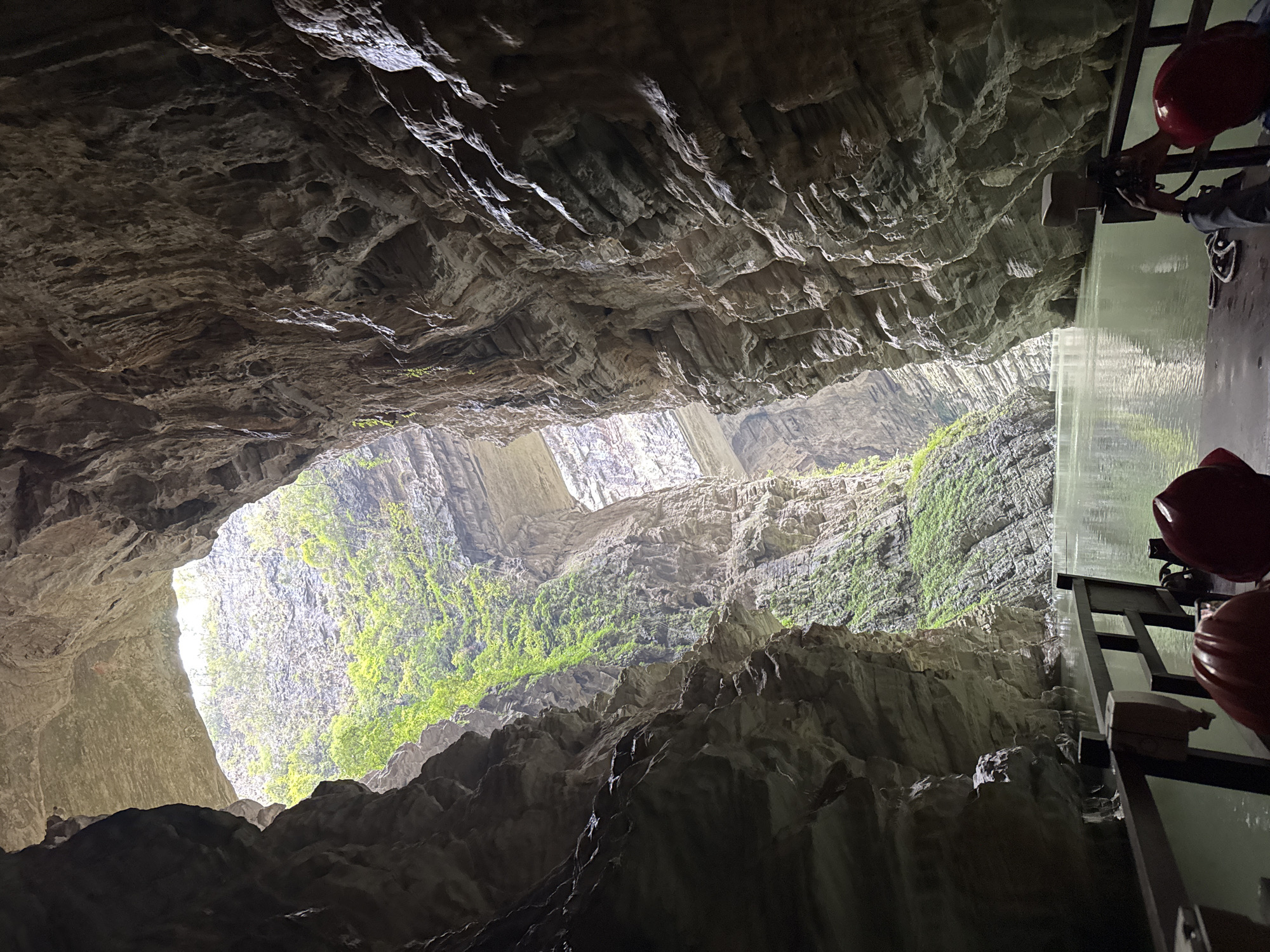 A low-angle, skewed perspective captures a deep cavern in Unknown, Unknown. The cavern walls, textured with layered rock formations, converge towards a central opening where sunlight streams through, illuminating patches of green vegetation clinging to the rock face. A man stands at the right, seemingly operating the structure in the background, wearing a red hard hat and with his hands touching what looks like the waterfall. The background shows what looks like the other side of the cavern with green vegetation.
