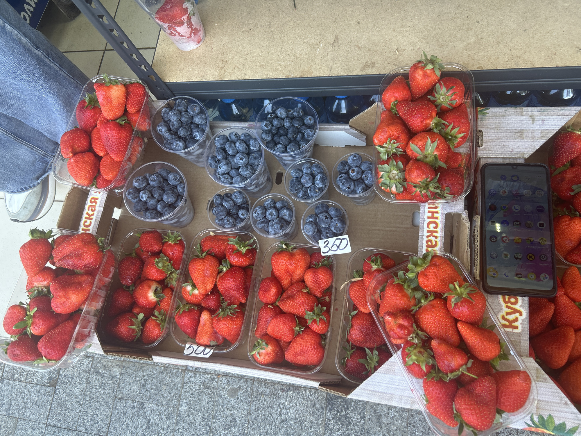 The image shows a street vendor's display of strawberries and blueberries in an unknown city. The fruit is displayed in clear plastic containers of various sizes. Strawberries are in rectangular containers, some with price tags of "500" and "350," while blueberries are in cylindrical cups. The backdrop features a partially visible person wearing jeans, and a mobile phone rests atop a strawberry container. The fruits are arranged on what appears to be a cardboard box, suggesting a makeshift vending setup.